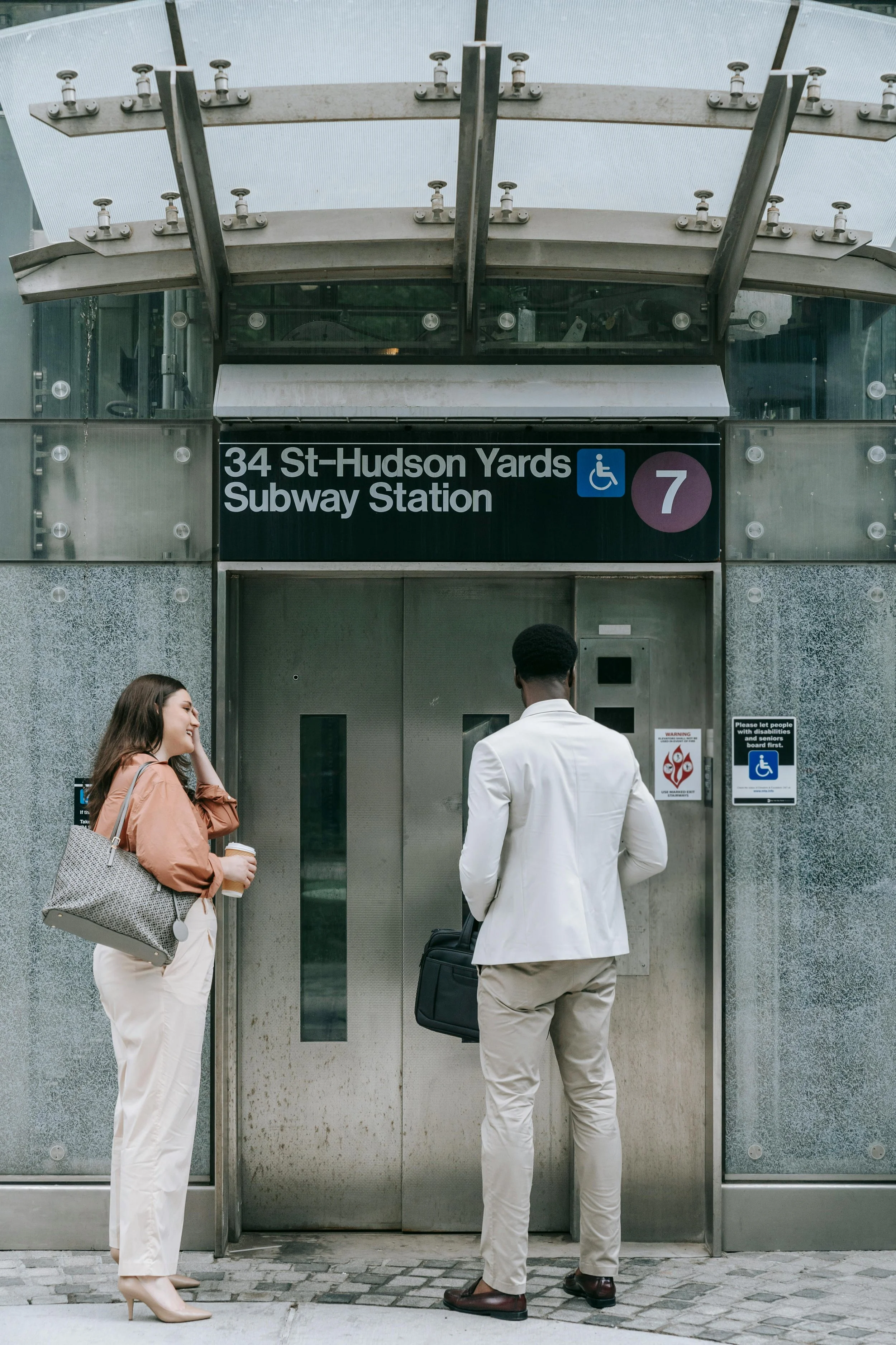 Two people stand outside the entrance to the 34th Street-Hudson Yards subway station in New York City. One woman is smiling, holding a coffee, and touching her hair. The other person, a man in a white blazer, is holding a black bag and facing the elevator doors.