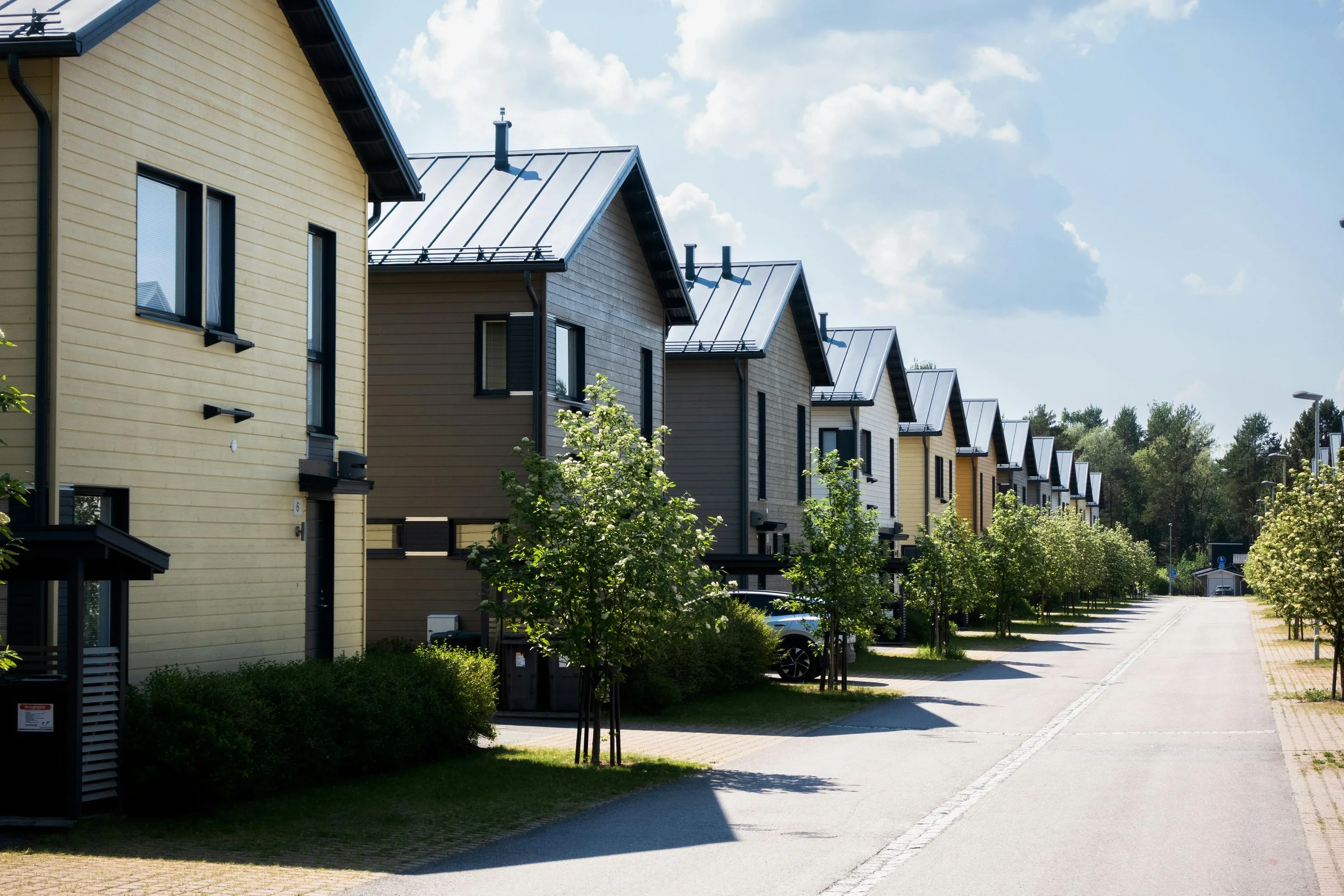 Row of modern, multicolored houses on a suburban street with trees and parked cars, under a partly cloudy sky.