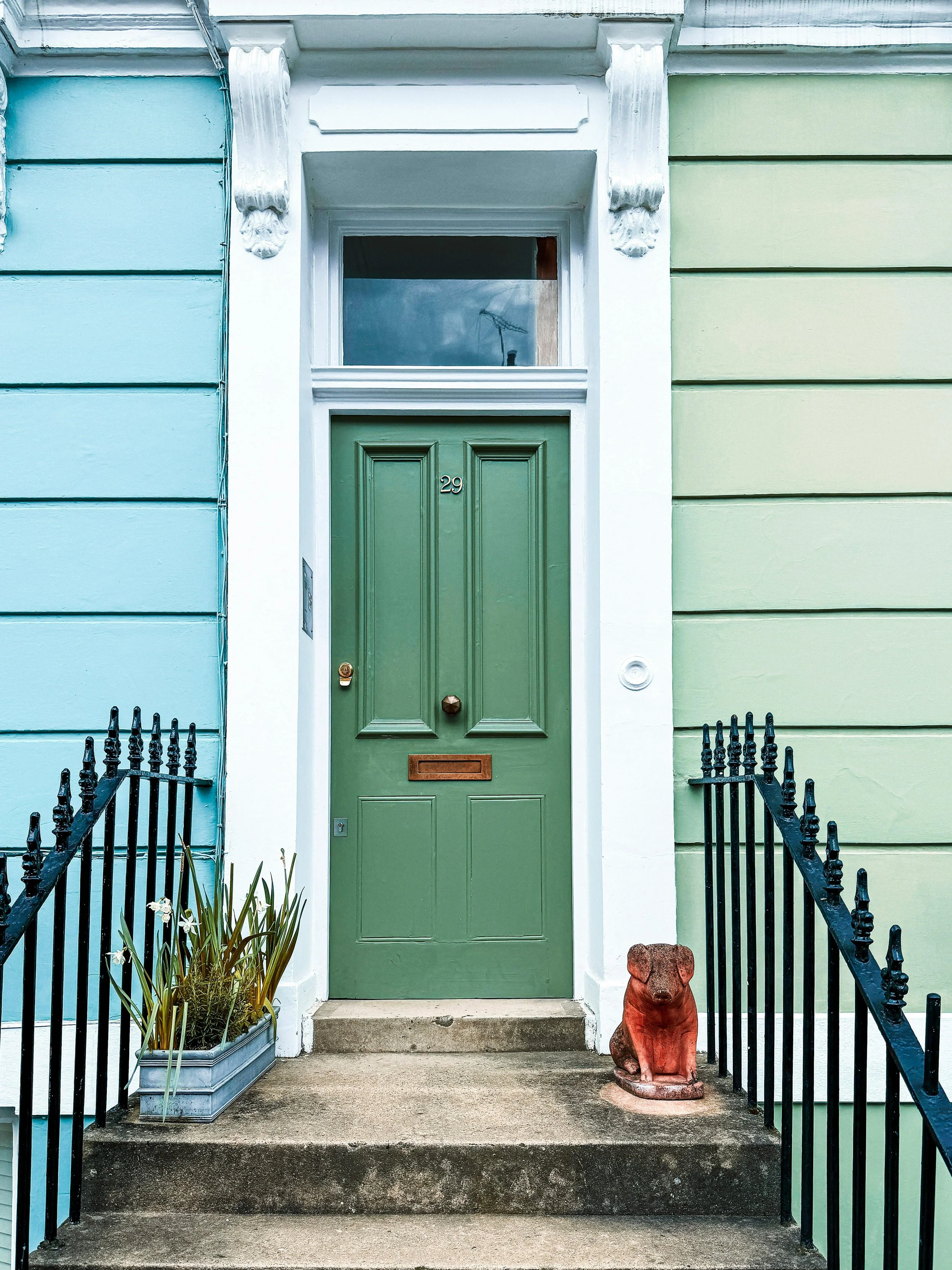 Green front door with the number 29, flanked by multicolored exterior walls, with black railings on stone steps, a planter with plants on the left, and a dog statue on the right.