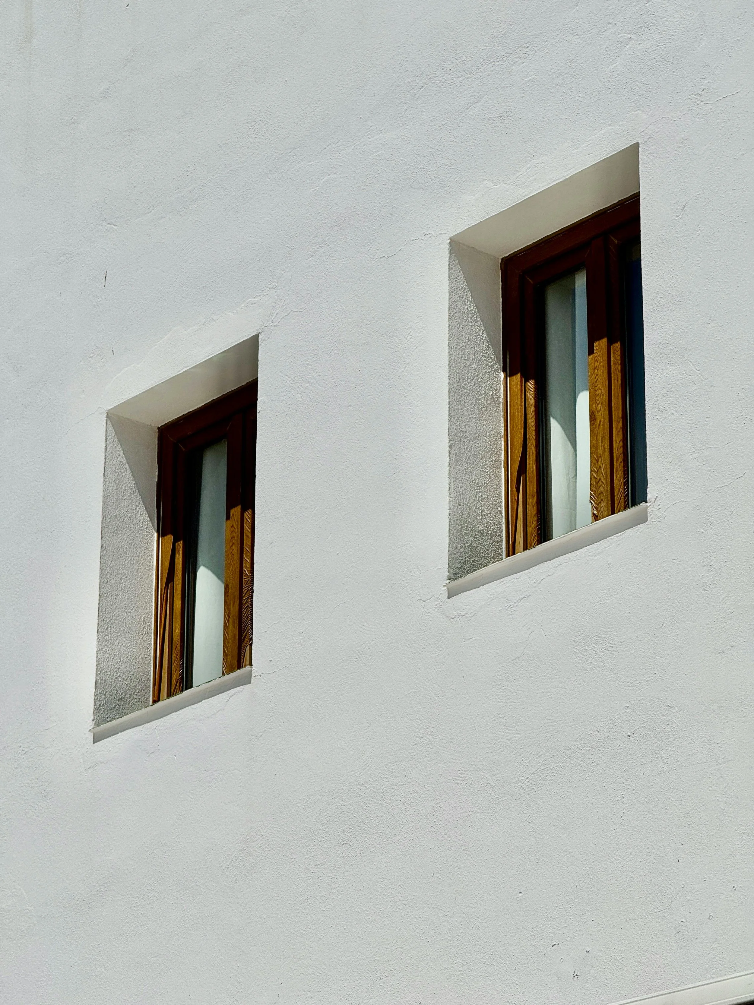 Two small windows with wooden frames on a white exterior wall, casting shadows.