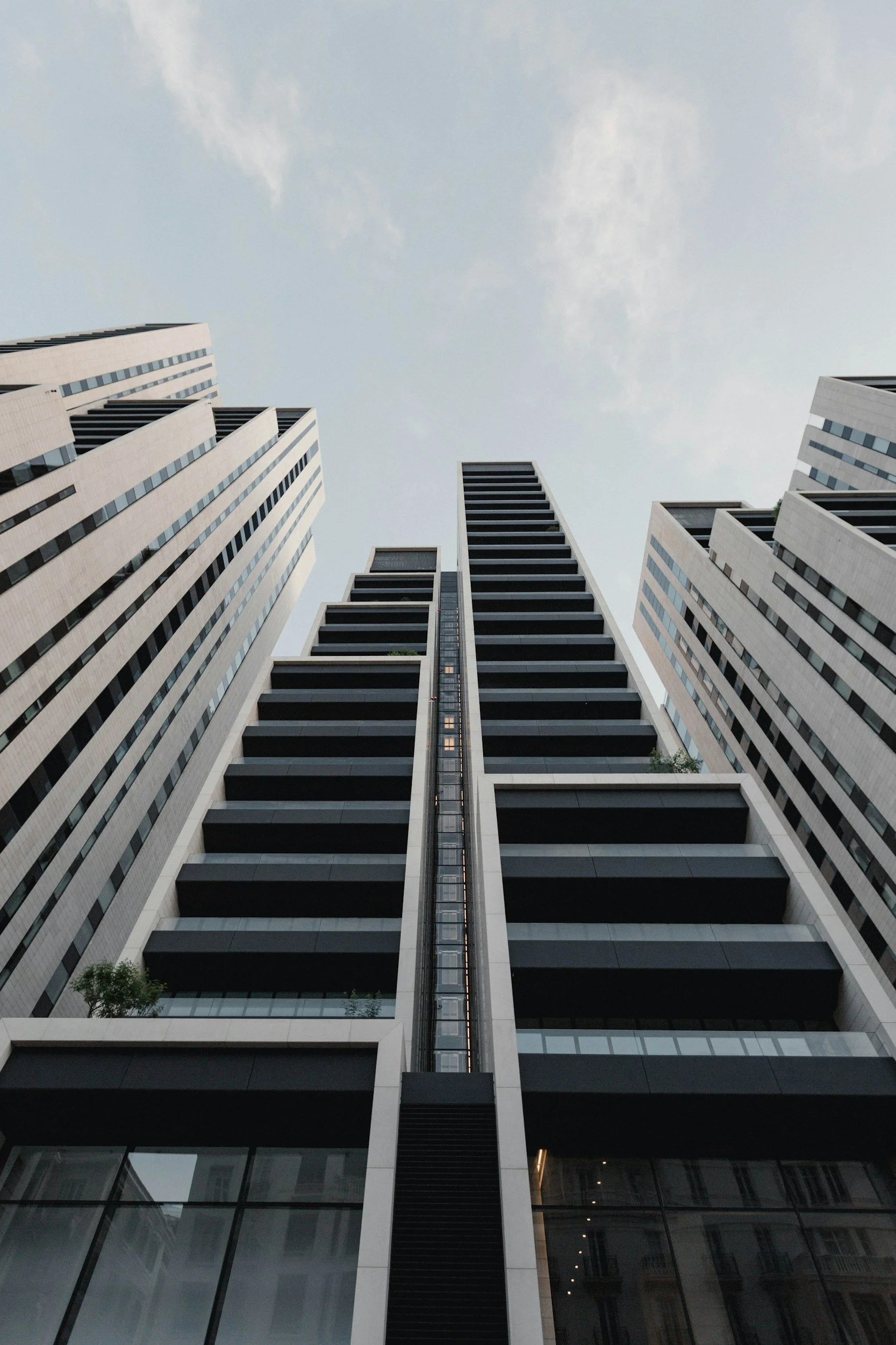 A view looking up at tall modern skyscrapers with glass and concrete facades, reaching towards a cloudy sky.