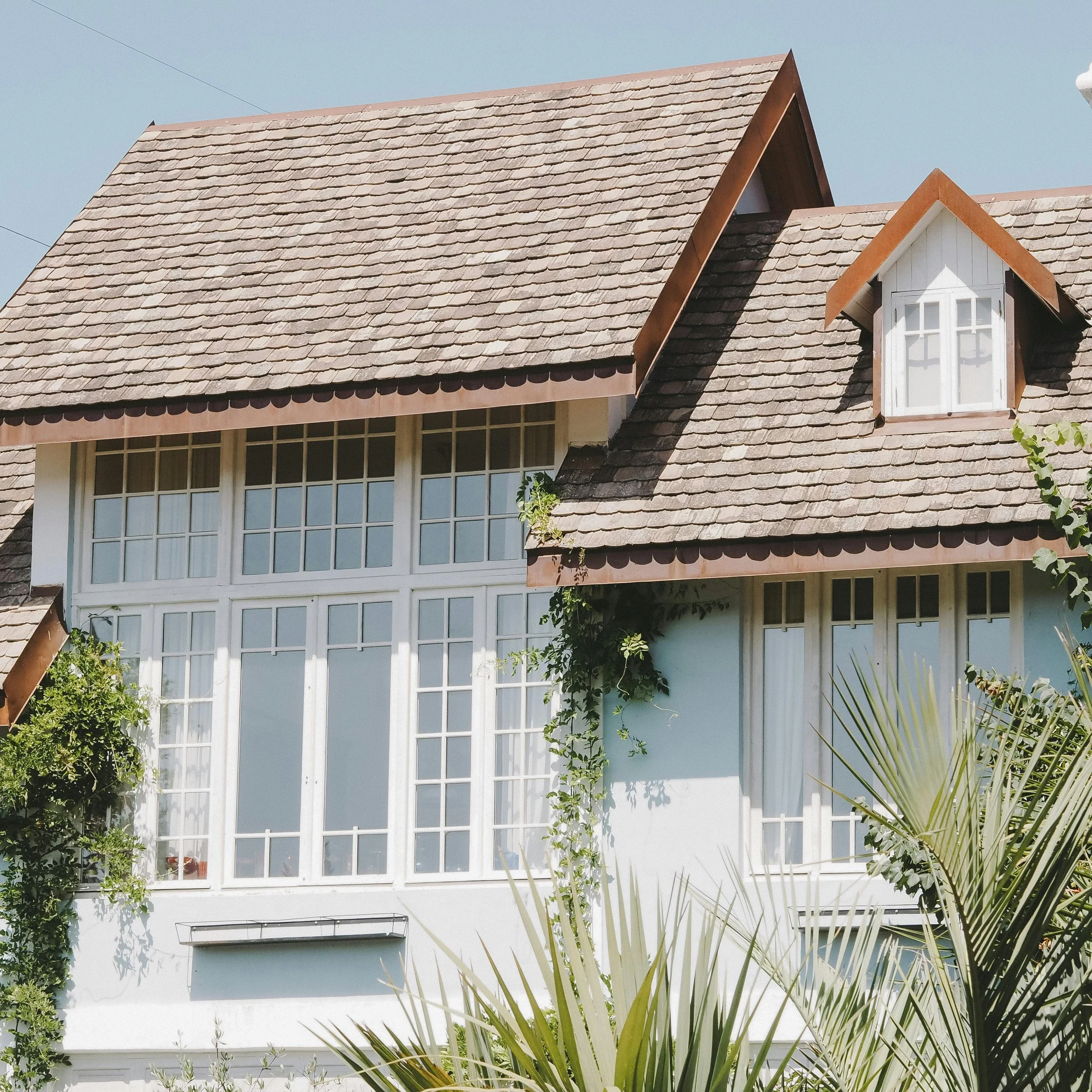 A house with multiple large windows, a shingled roof, and a small dormer window, partially covered by greenery and palm leaves.