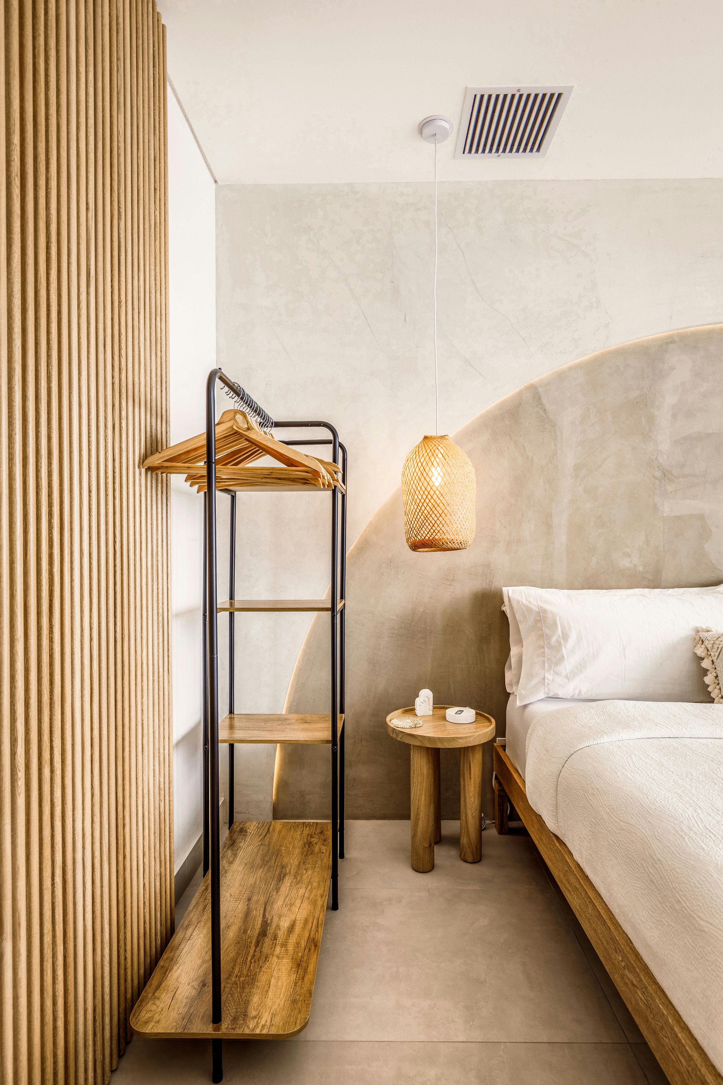 A cozy bedroom corner featuring a wooden bed frame with white bedding, a small round wooden side table with an alarm clock, a hanging woven pendant light, a black metal and wood clothing rack with wooden hangers, and a beige textured wall with a curved design.