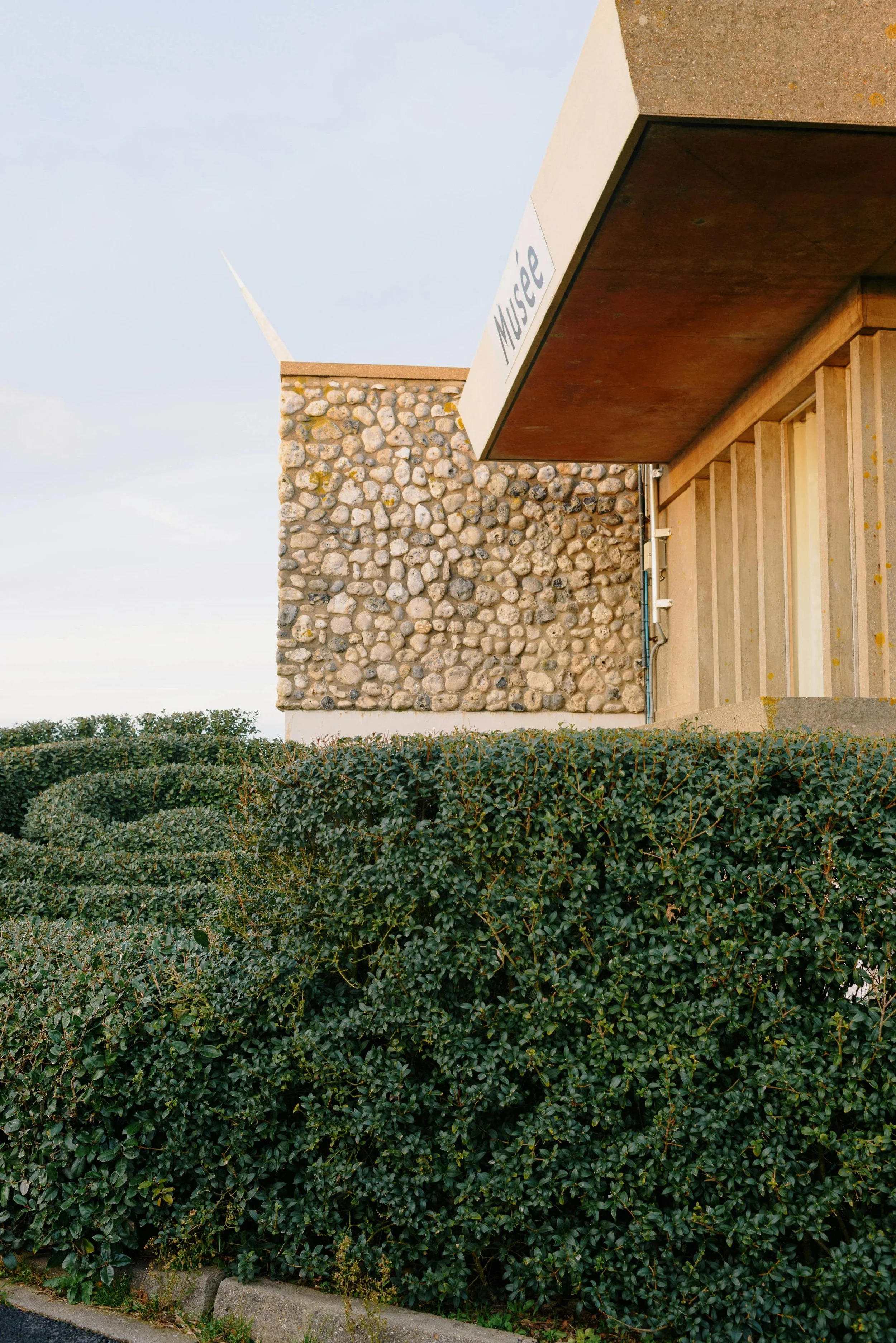 View of a museum building with a textured stone wall, a sign that reads 'Museum', and a neatly trimmed green shrub in front.