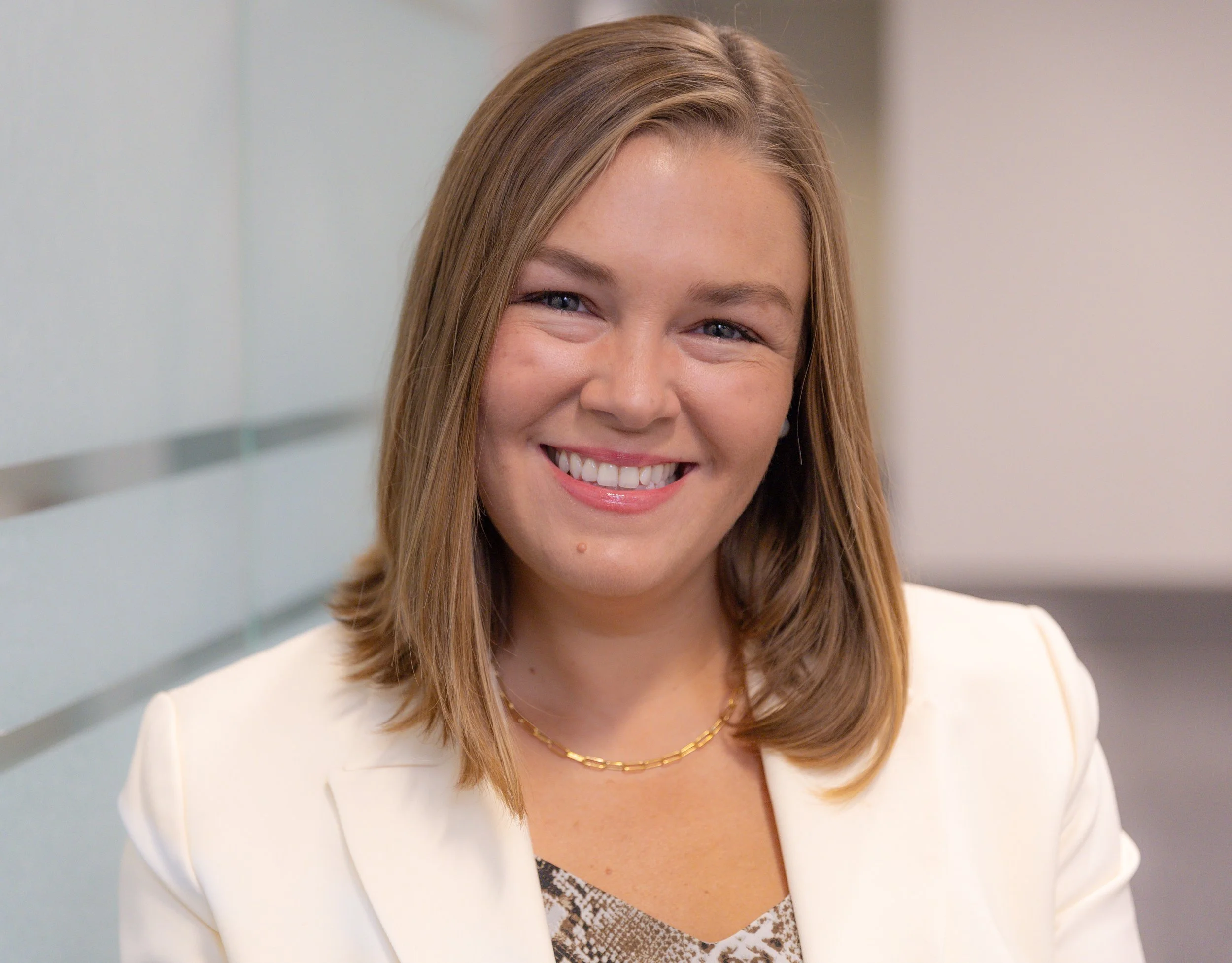 A woman with shoulder-length light brown hair smiling, wearing a white blazer, a gold chain necklace, and a patterned top, standing in front of a glass wall.