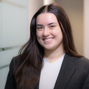 Portrait of a woman with long dark hair smiling, wearing a dark blazer and white top, in an indoor office setting.