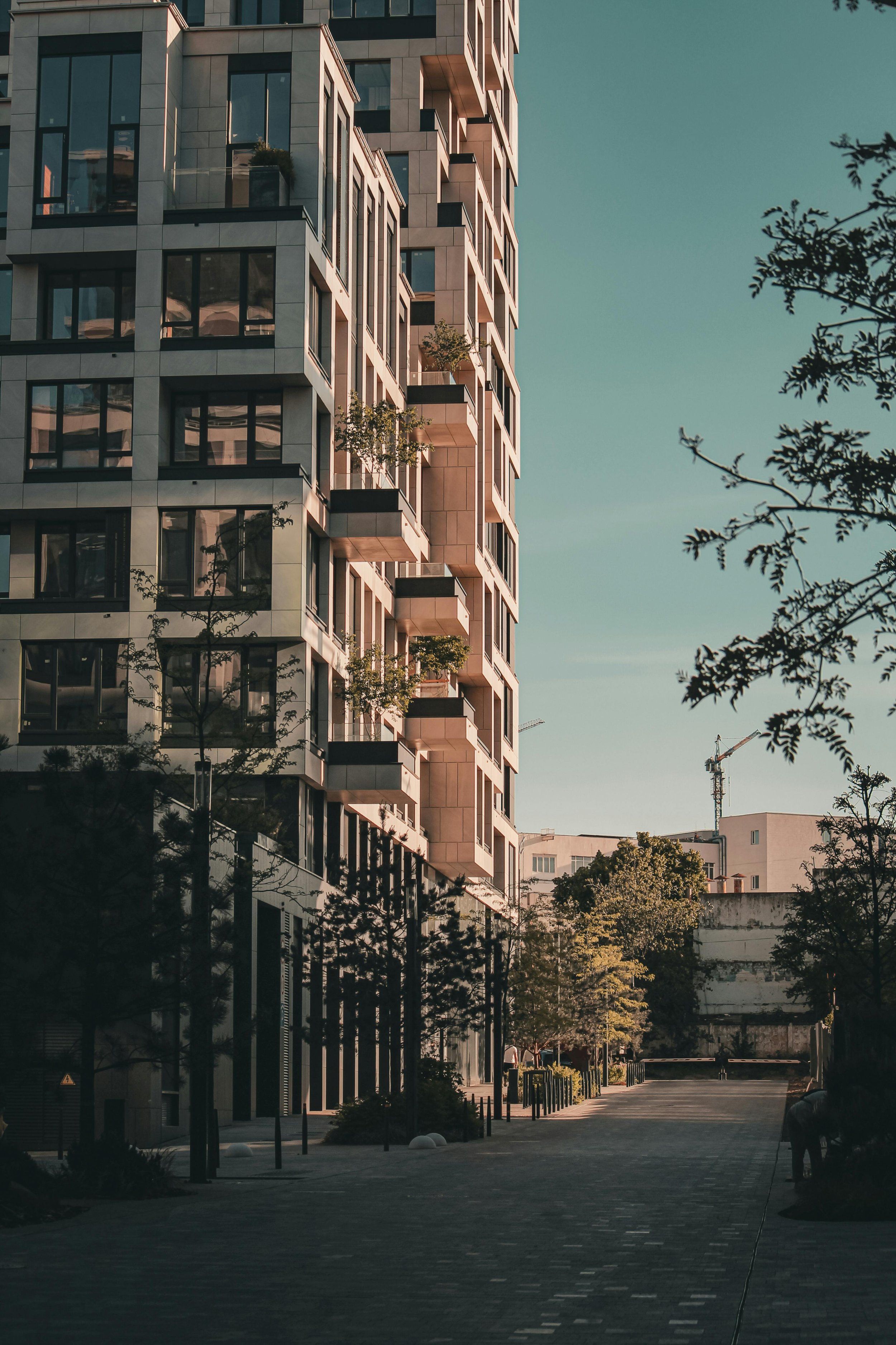 A modern multi-story apartment building with large glass windows and small balconies, situated along a paved pedestrian street lined with trees and bushes. The sky is clear with a few clouds and there are construction cranes in the background.