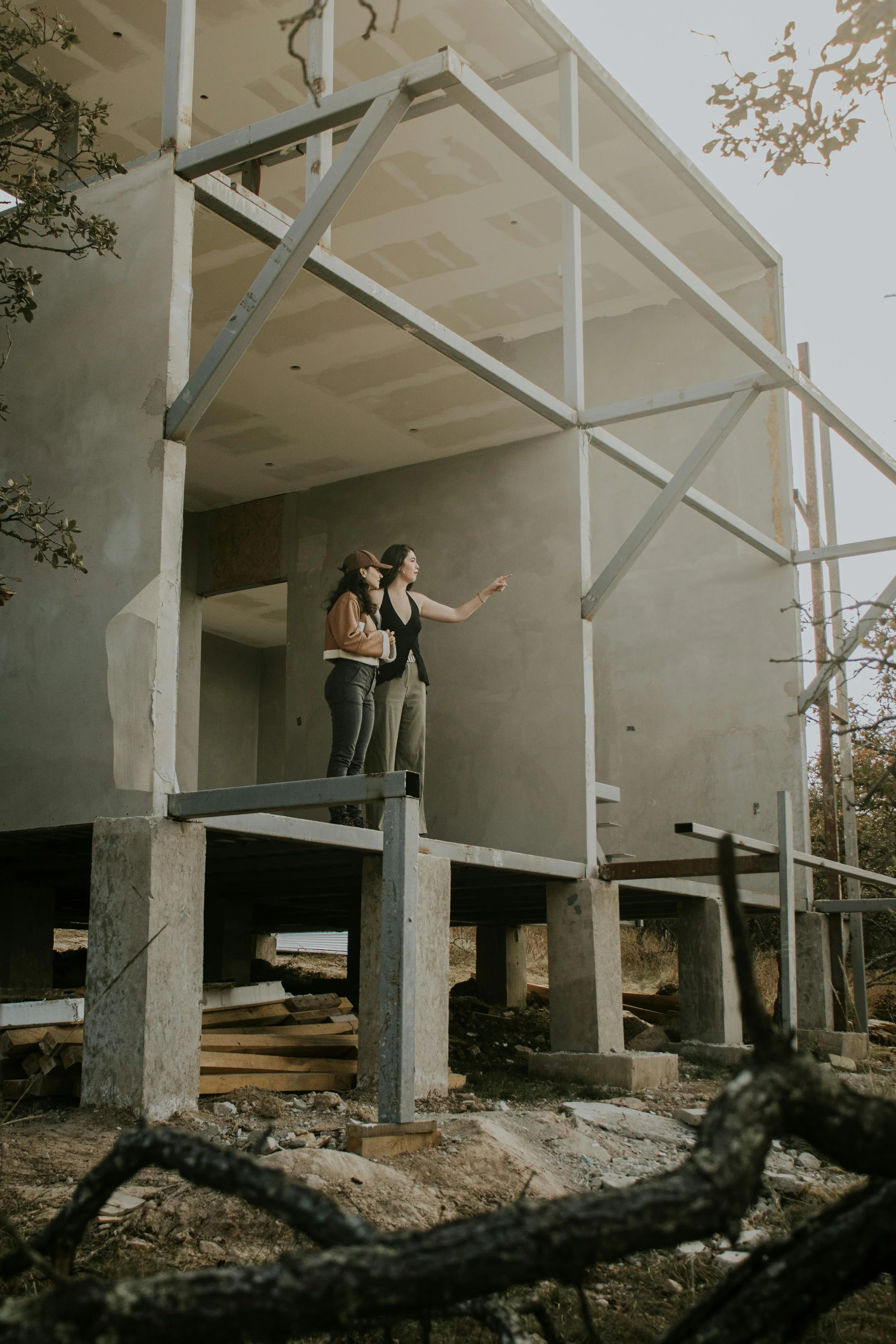 Two women stand on a construction site, examining a partially built house. One woman points towards the house while the other listens. The house is elevated on stilts, with exposed framework and construction materials visible below.