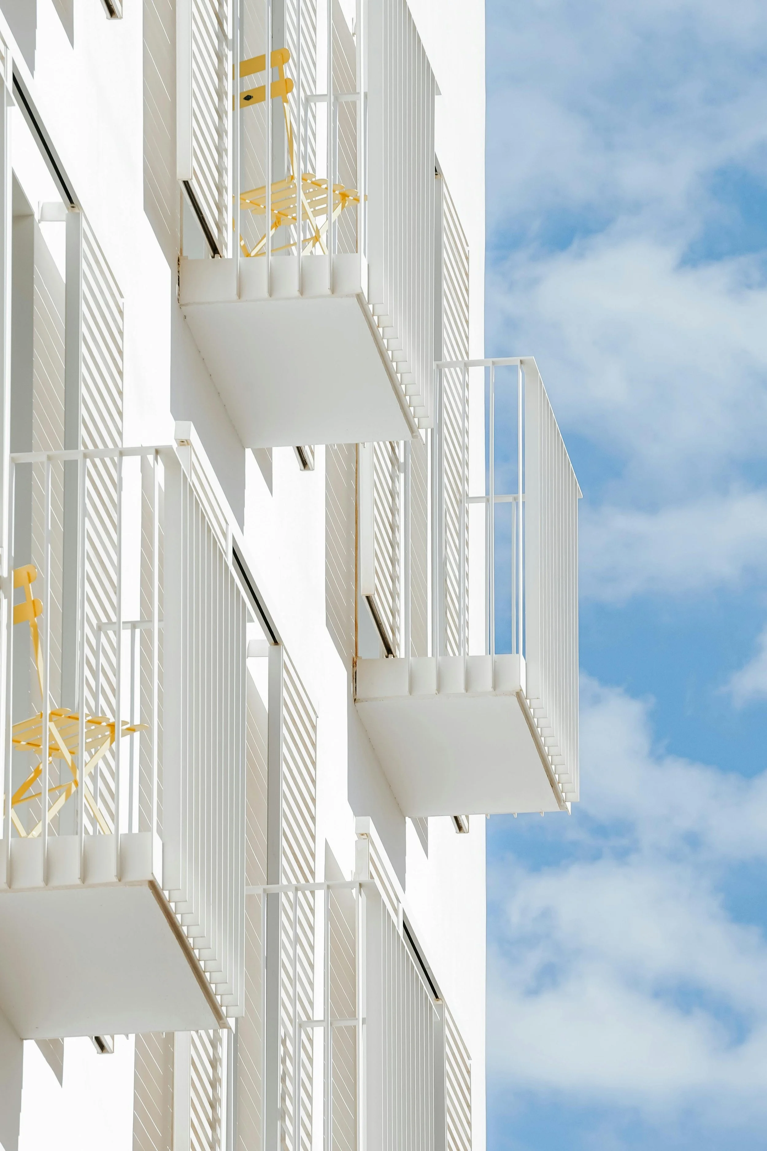 Multiple white balconies on a white building with yellow chairs, against a blue sky with clouds.