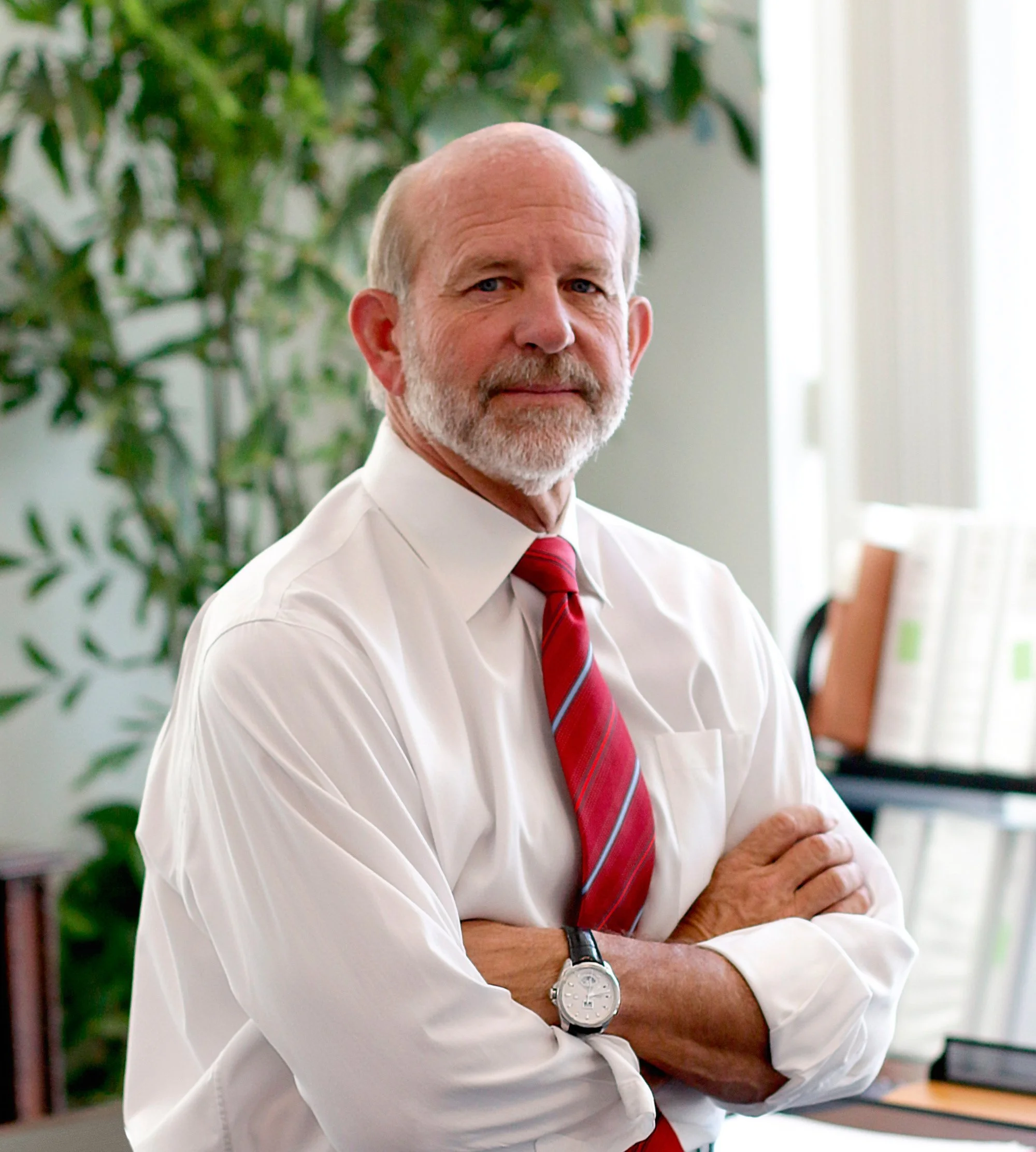 An older man with a beard and mustache, wearing a white dress shirt, red striped tie, and a wristwatch, standing with arms crossed in an office setting.