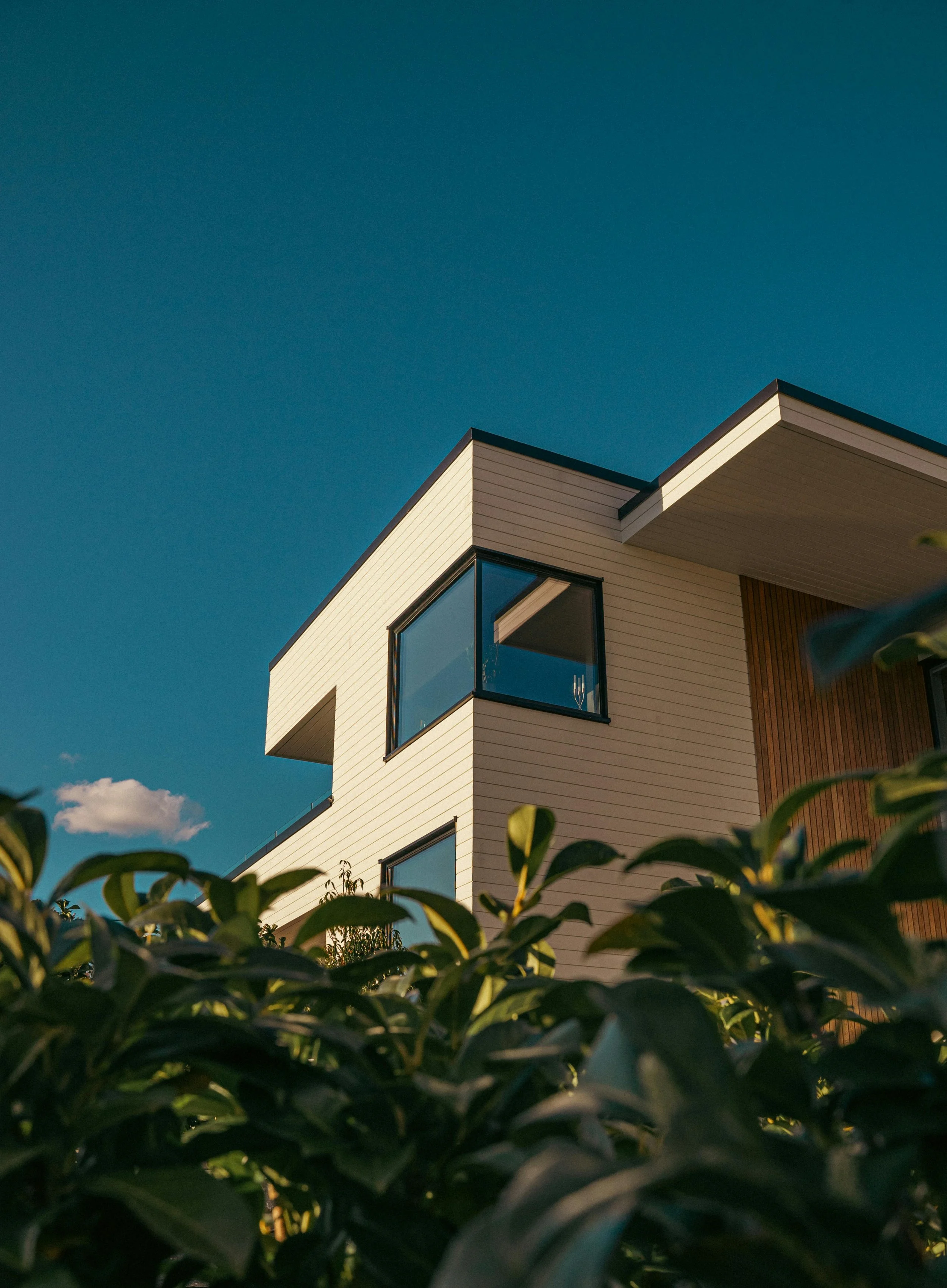 Modern two-story house with large windows, white siding, and wooden accents, photographed from a low angle during daytime, with green foliage in the foreground and a clear blue sky in the background.