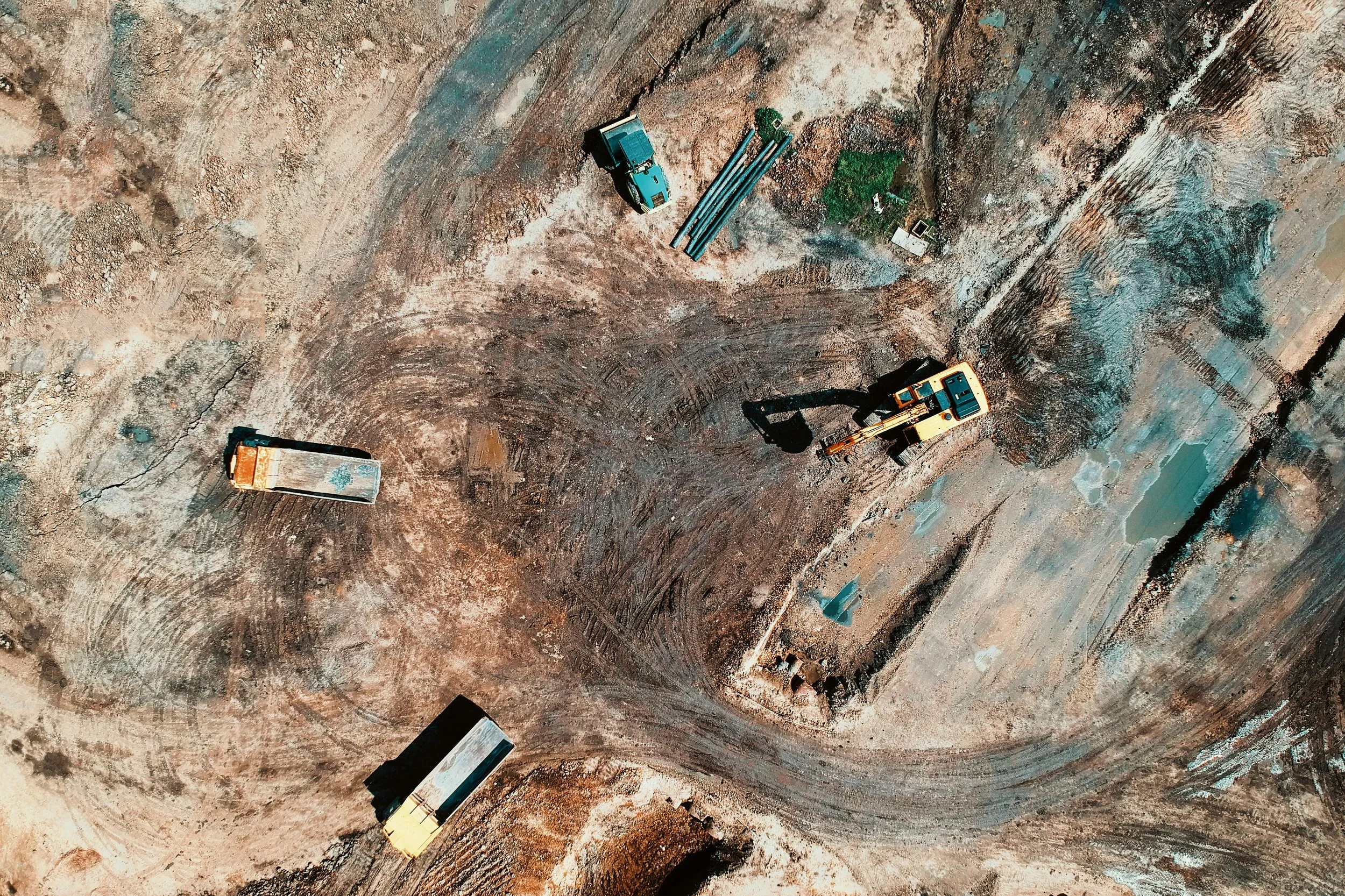 An aerial view of a construction site with heavy machinery and scattered materials, including two dump trucks, a small excavator, and construction pipes.
