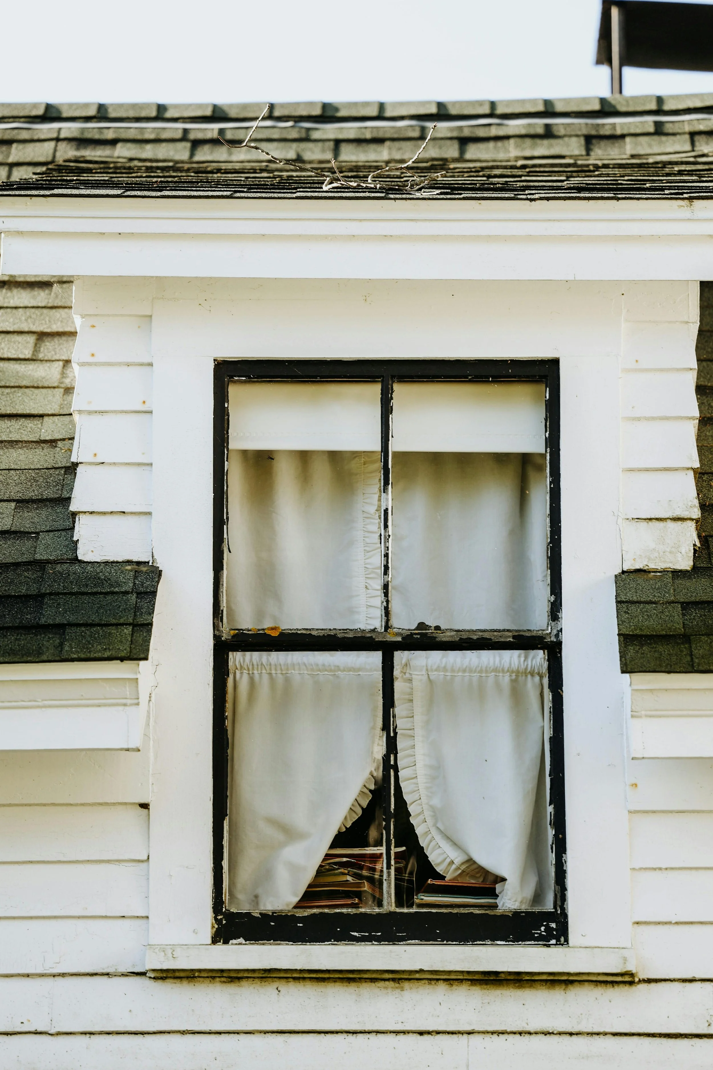 Old wooden house window with white curtains, some books visible inside, black-painted window frame, part of house siding and roof shingles visible.