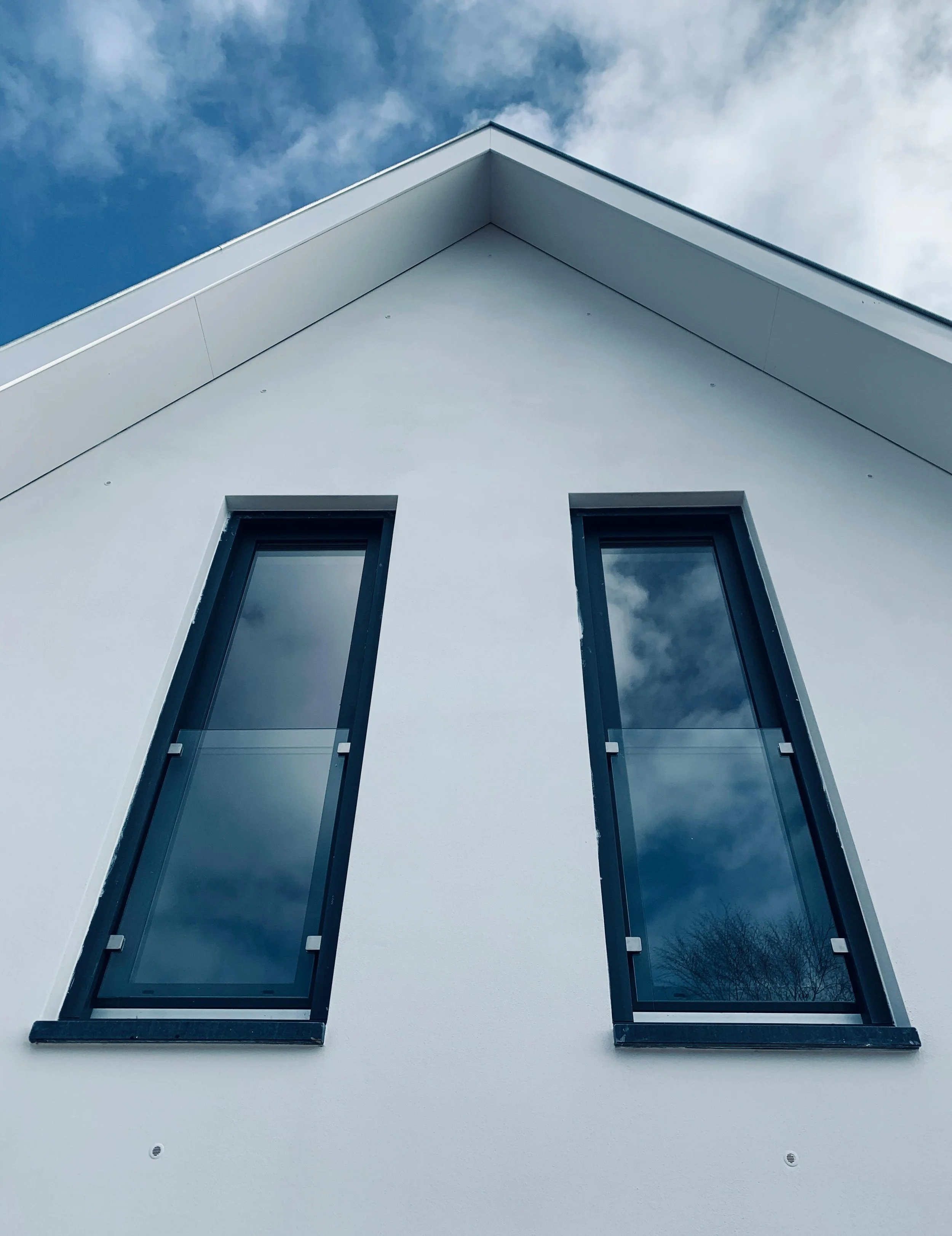 Modern white building with two large narrow windows reflecting a cloudy sky, viewed from below.