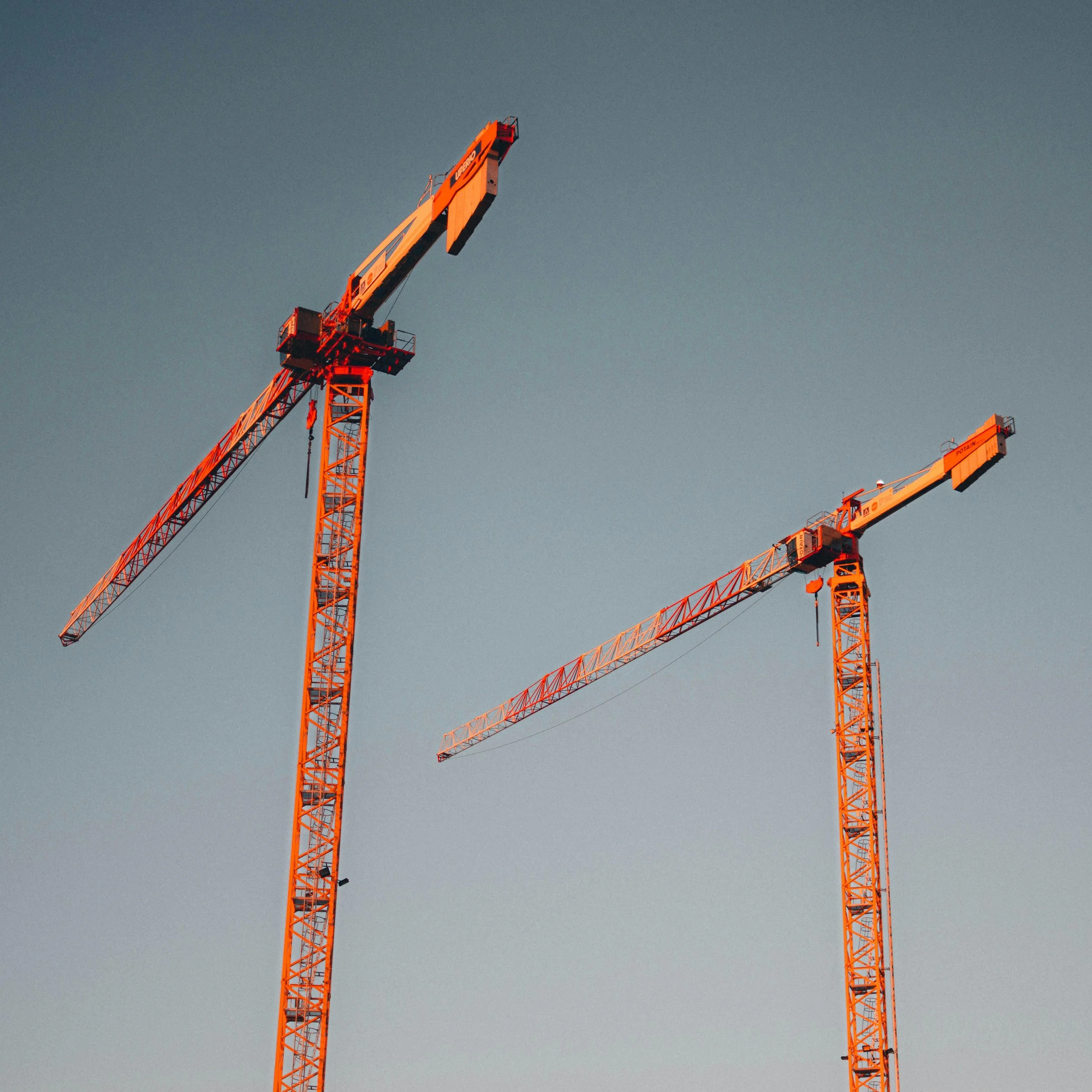 Two tall construction cranes with orange and white structural components against a dusk sky.