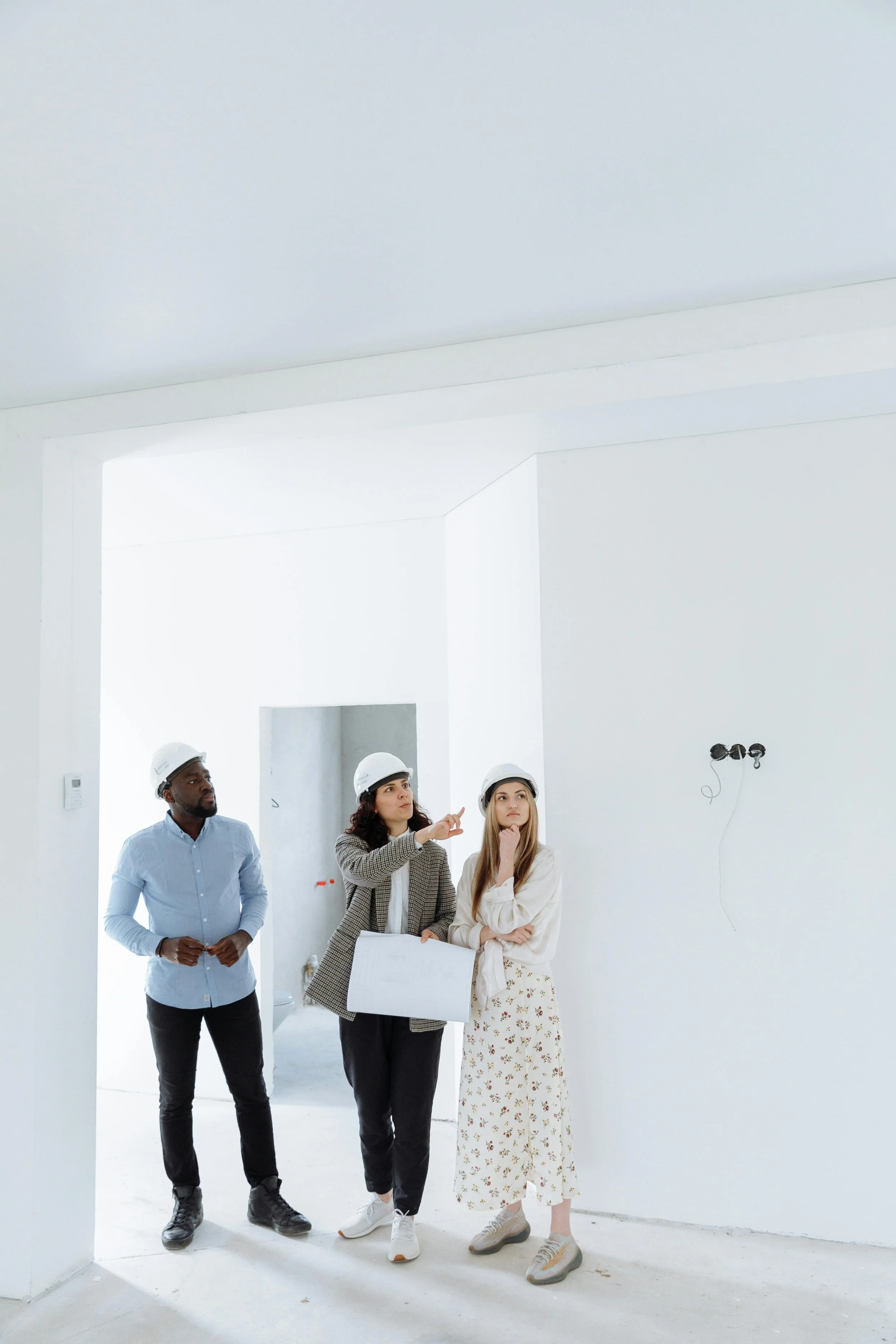 Three people wearing white construction helmets inside a building under construction, looking at the walls and discussing the project.