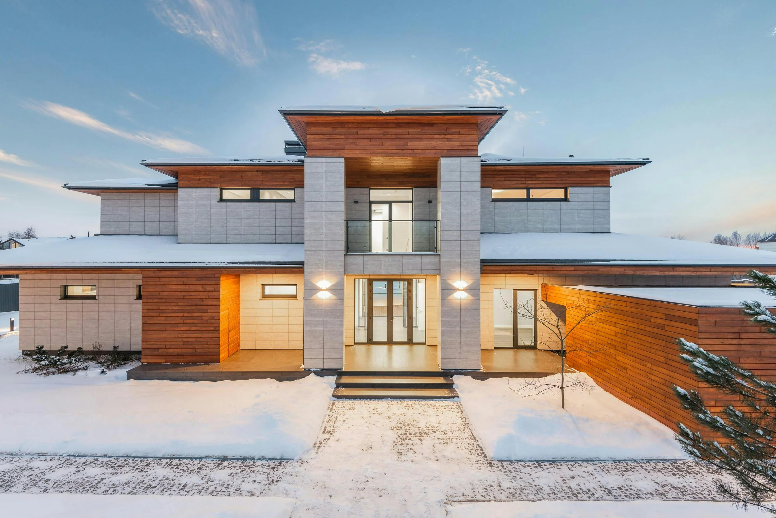 Modern multi-story house with gray stone and wooden exterior, snow-covered ground, and warm lighting inside and outside, during twilight.