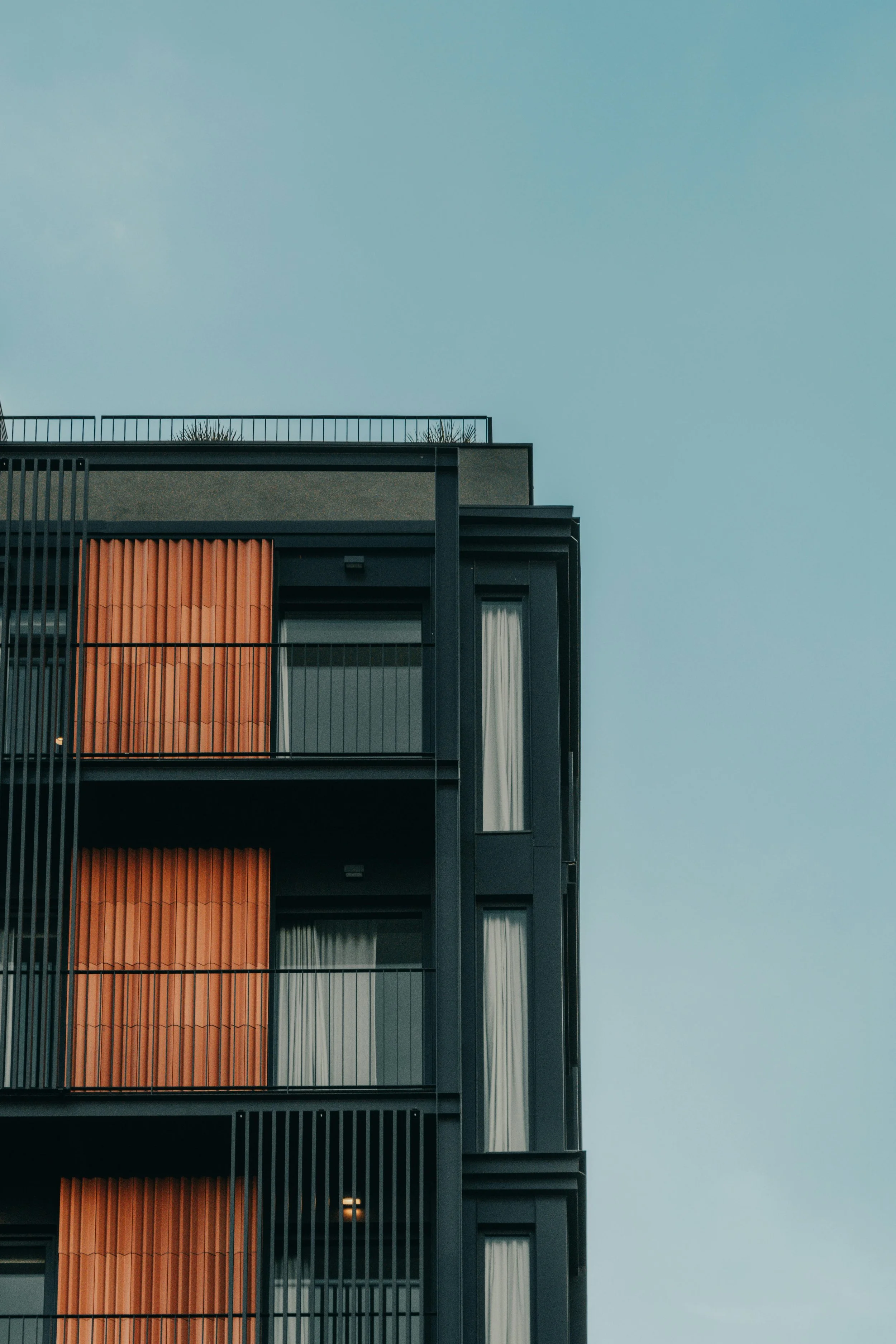Close-up of a modern apartment building with dark framing, balconies with black metal railings, orange vertical blinds, and white curtains, set against a clear blue sky.