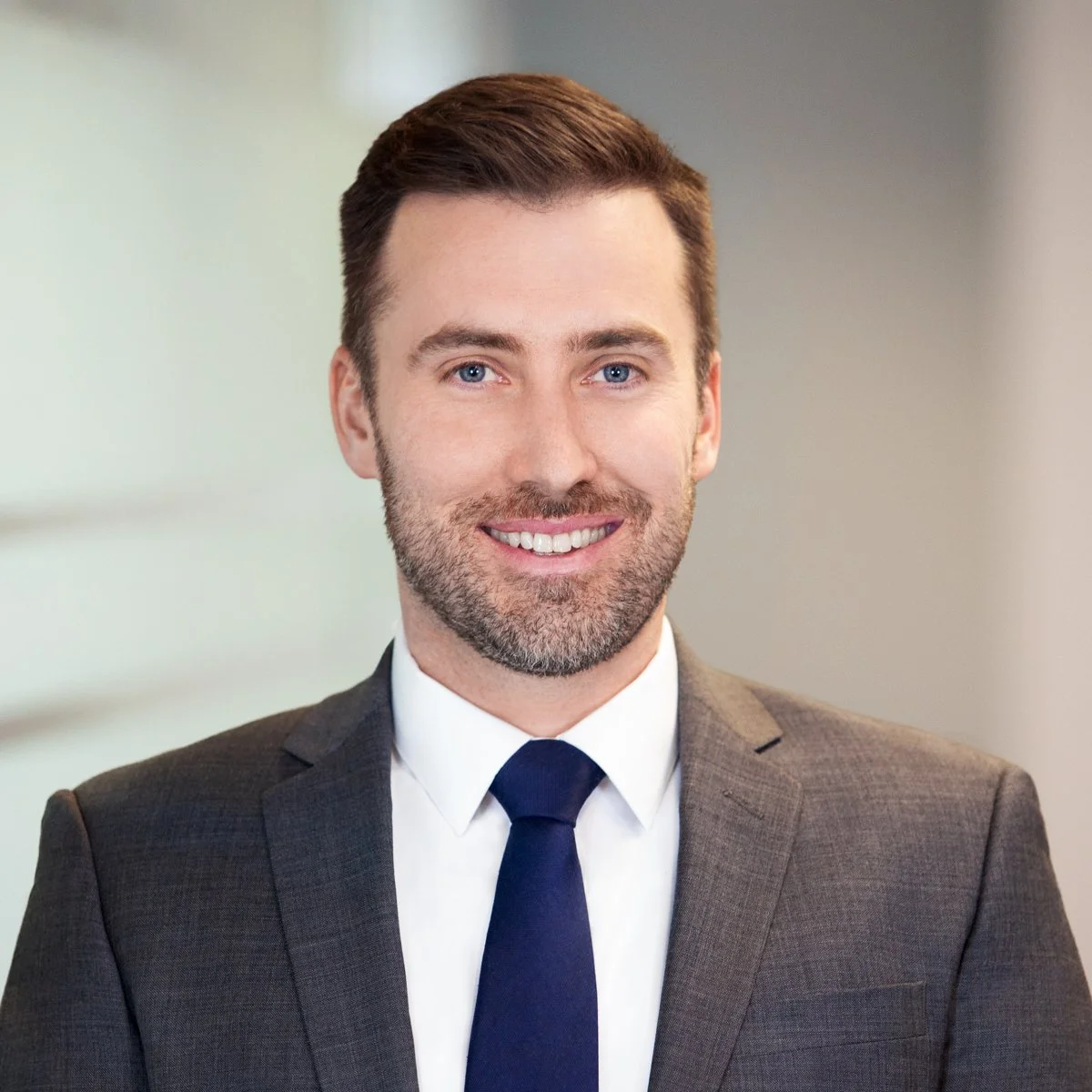 Portrait of a man with dark brown hair, blue eyes, and a beard, dressed in a gray suit with a white shirt and dark tie, smiling in an indoor setting.