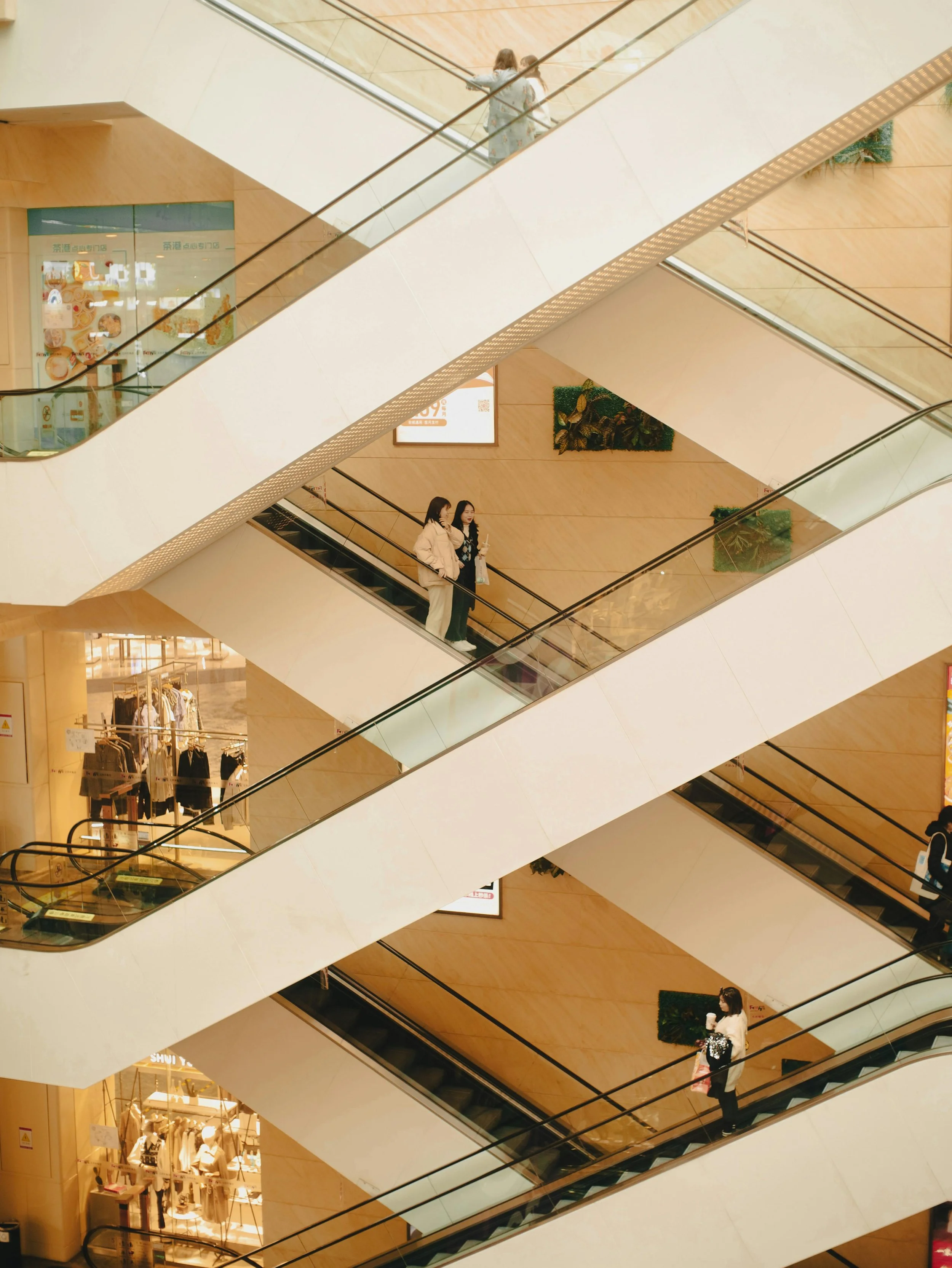 Multiple people riding escalators inside a shopping mall, with stores visible on either side, beige and wooden interior design, and some greenery decoration on the walls.