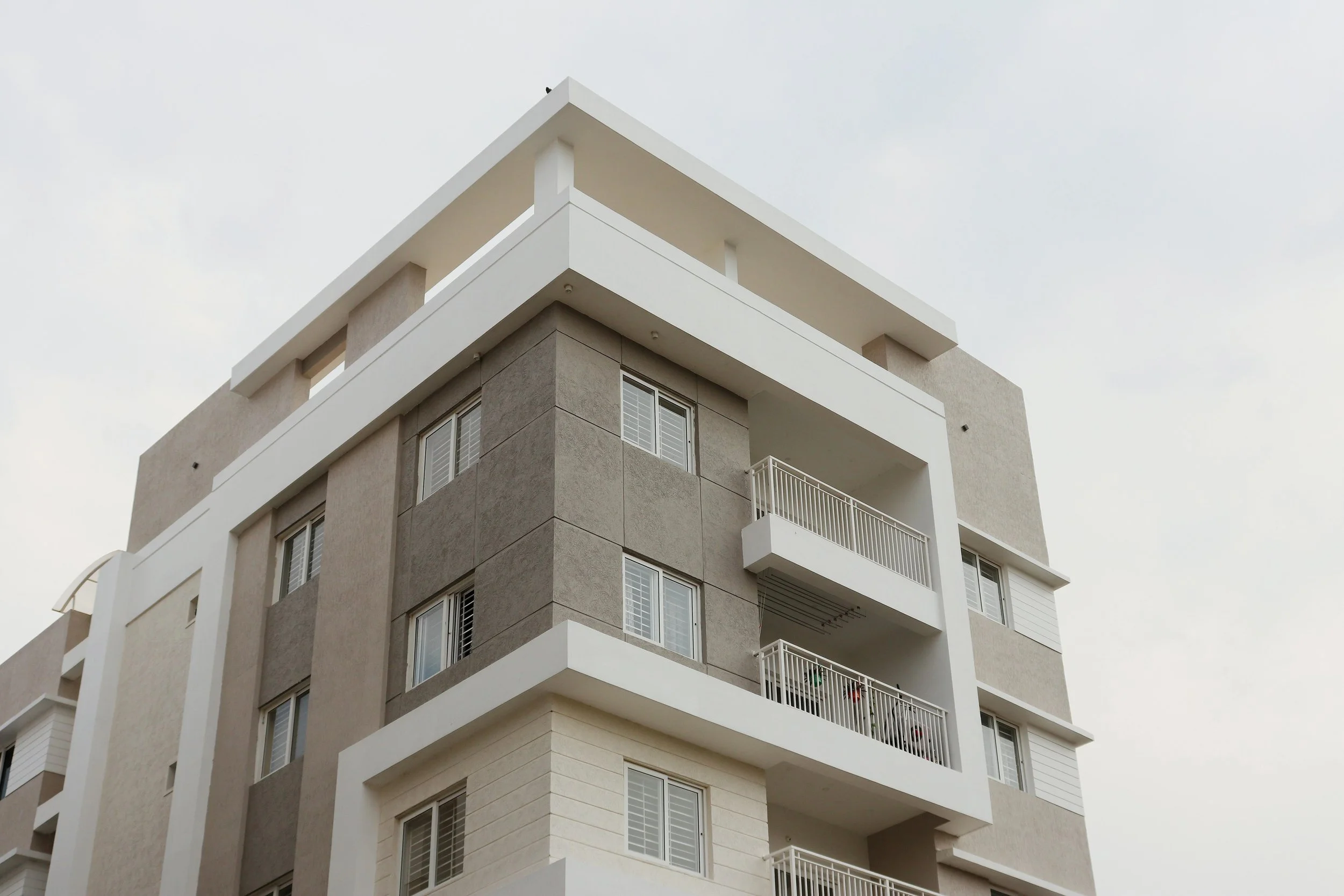 A modern multi-story residential building with beige and white exterior walls, several balconies with white railings, and windows with white shutters. The sky is overcast.