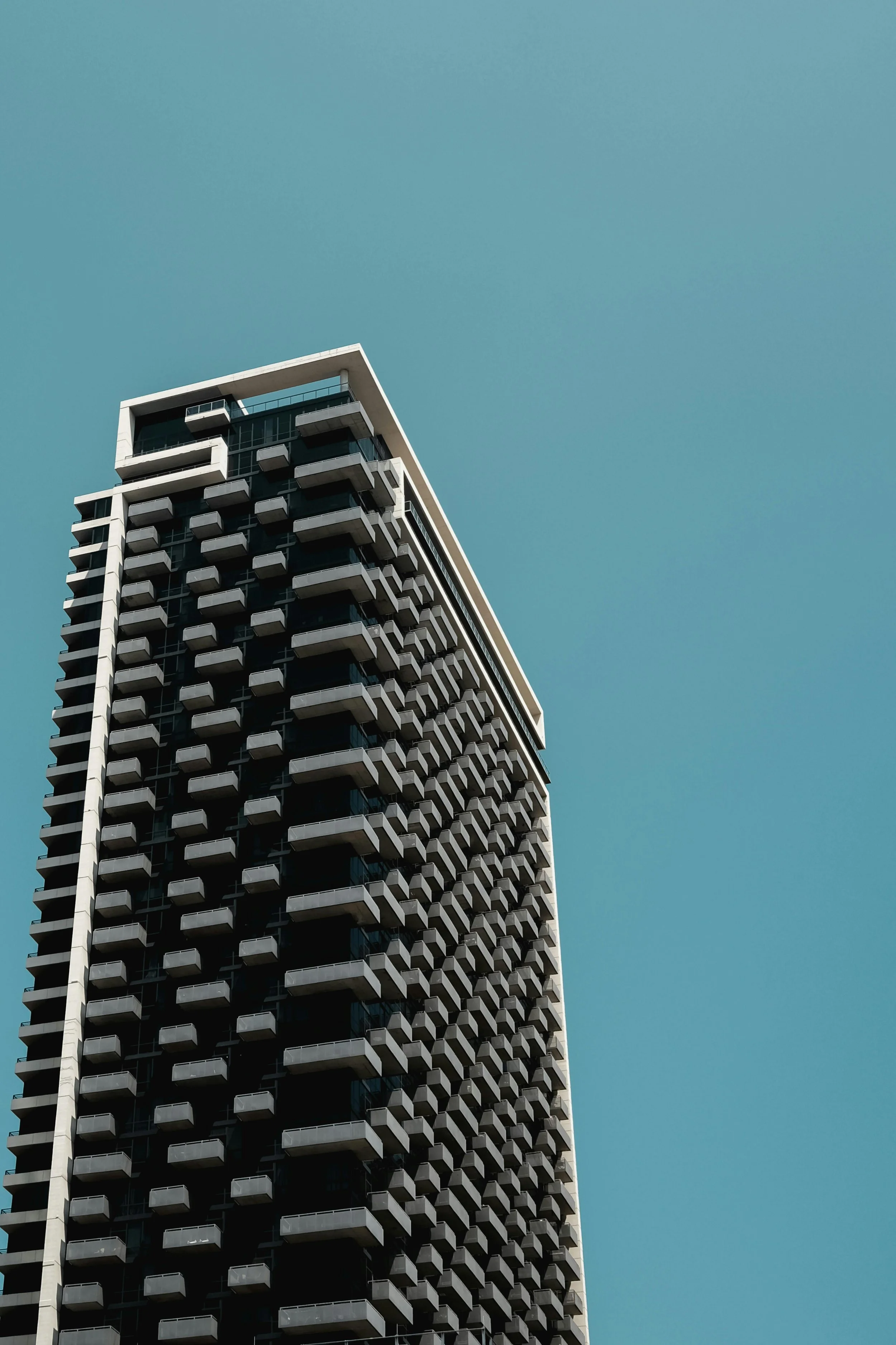 A modern high-rise building with multiple balconies, viewed from a low angle against a clear blue sky.