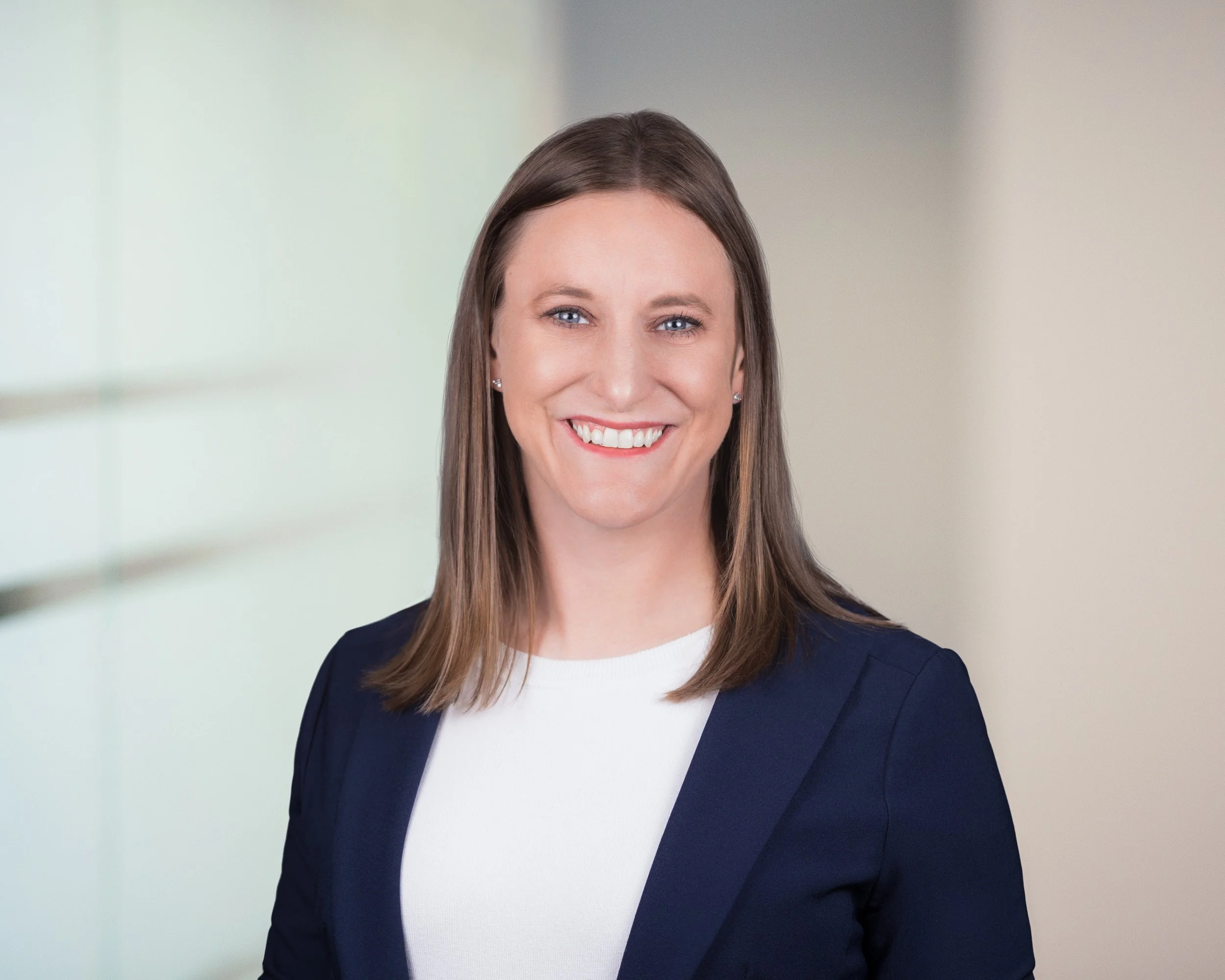 A smiling woman with straight, shoulder-length brown hair, wearing a white top and navy blazer, standing in front of a neutral background.