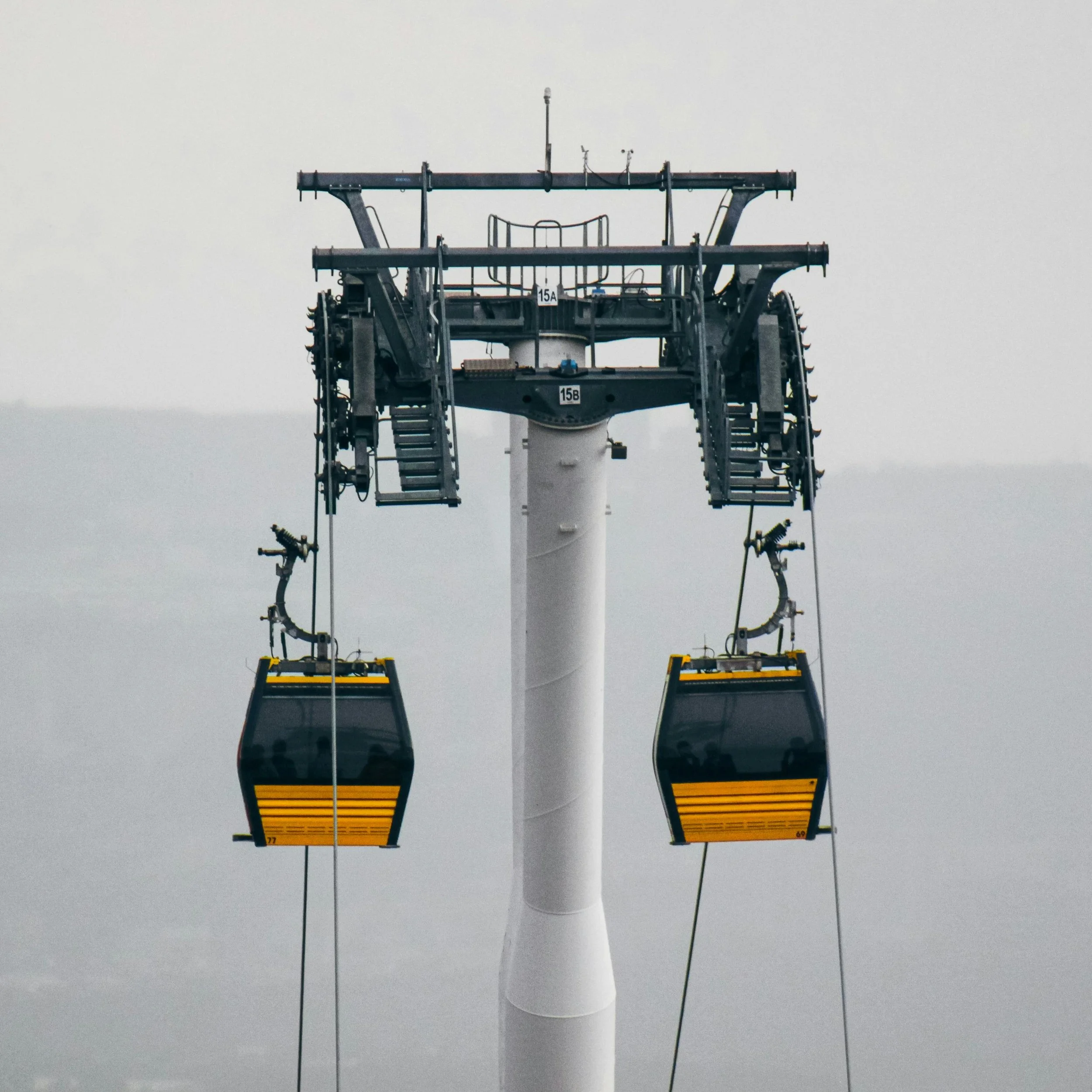 Close-up of a cable car tower with two yellow and black passenger cabins suspended from cables against a cloudy sky.