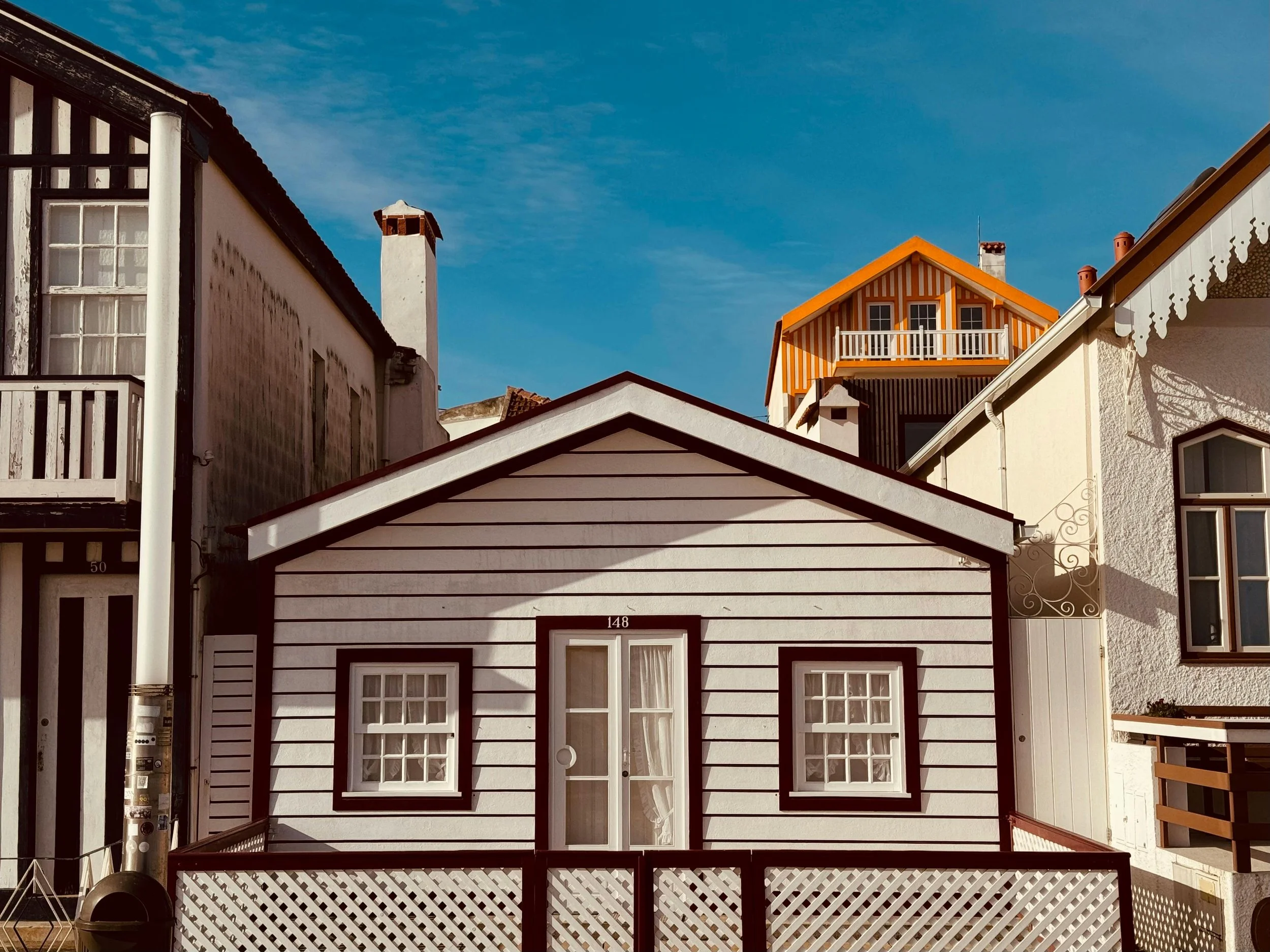 A small white house with black trim, three windows, and a door, situated between taller houses, under a clear blue sky.