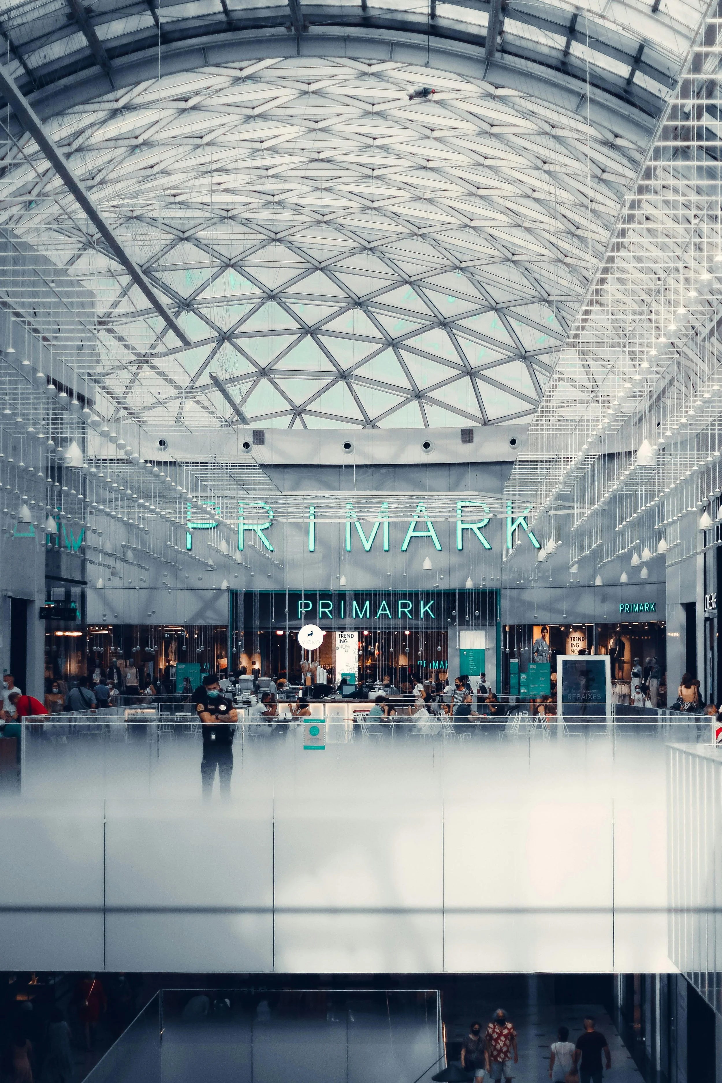 Interior view of a modern shopping mall with a large glass-domed ceiling, featuring a Primark store at the upper level, with shoppers and staff inside.