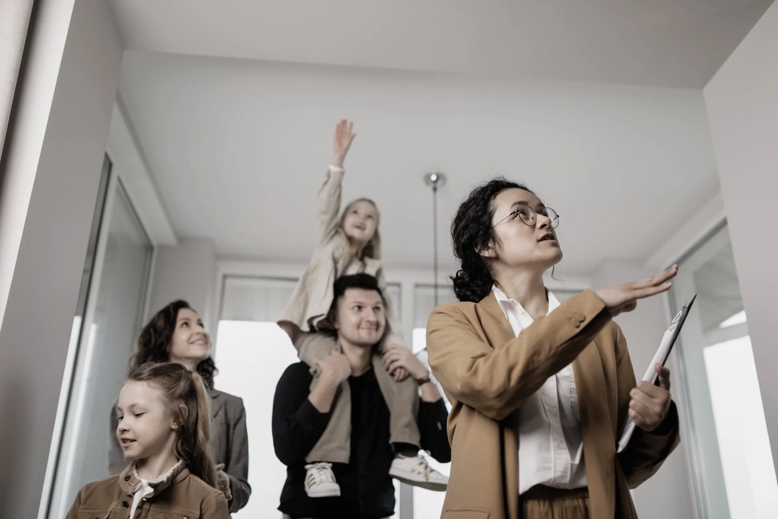 A group of adults and children inside an apartment, with some of the kids sitting on a man's shoulders, while a woman points at something and others watch or participate in a conversation.