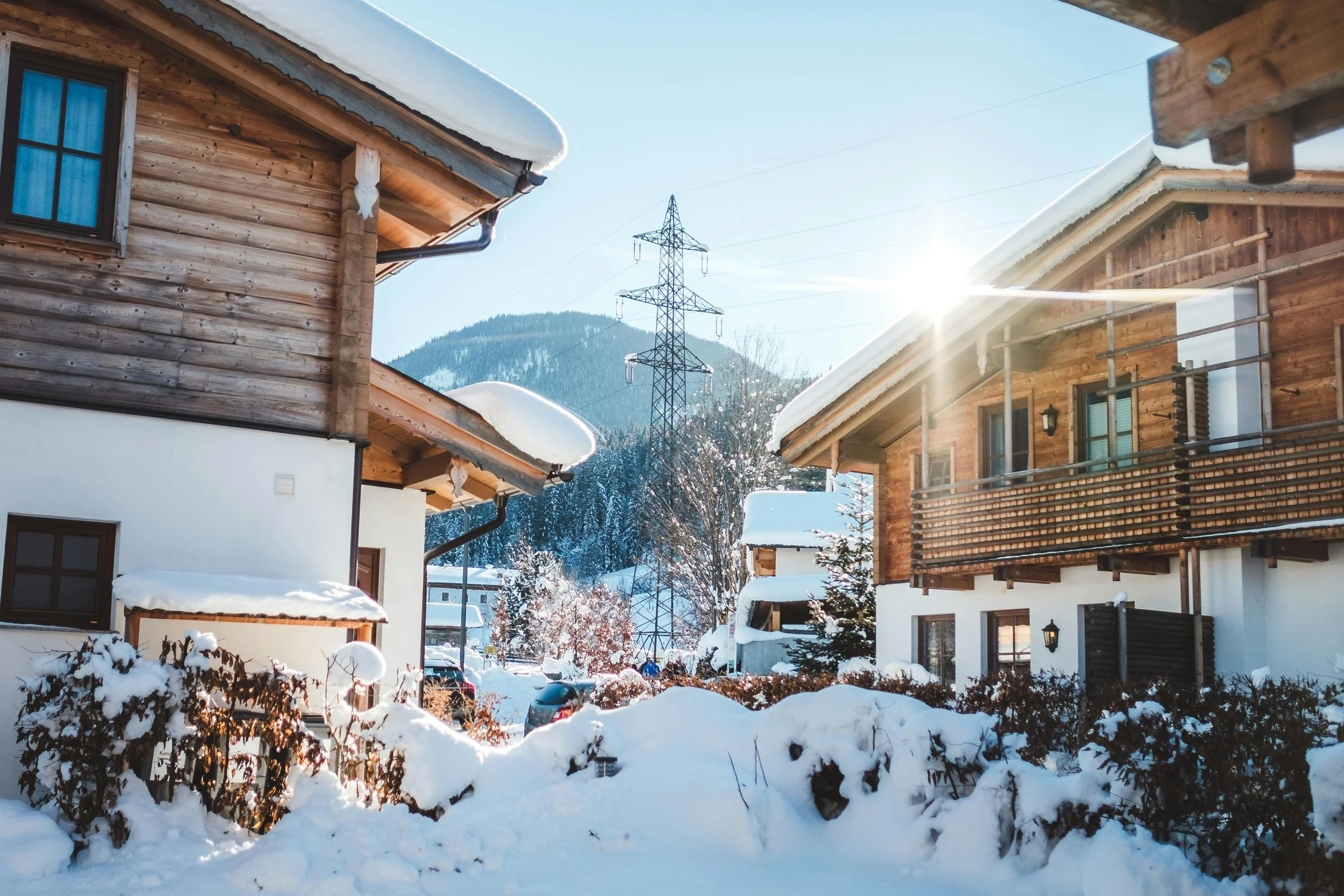 Snow-covered wooden houses in a mountain village with a power line and a mountain in the background, sunlight shining through clear blue sky.