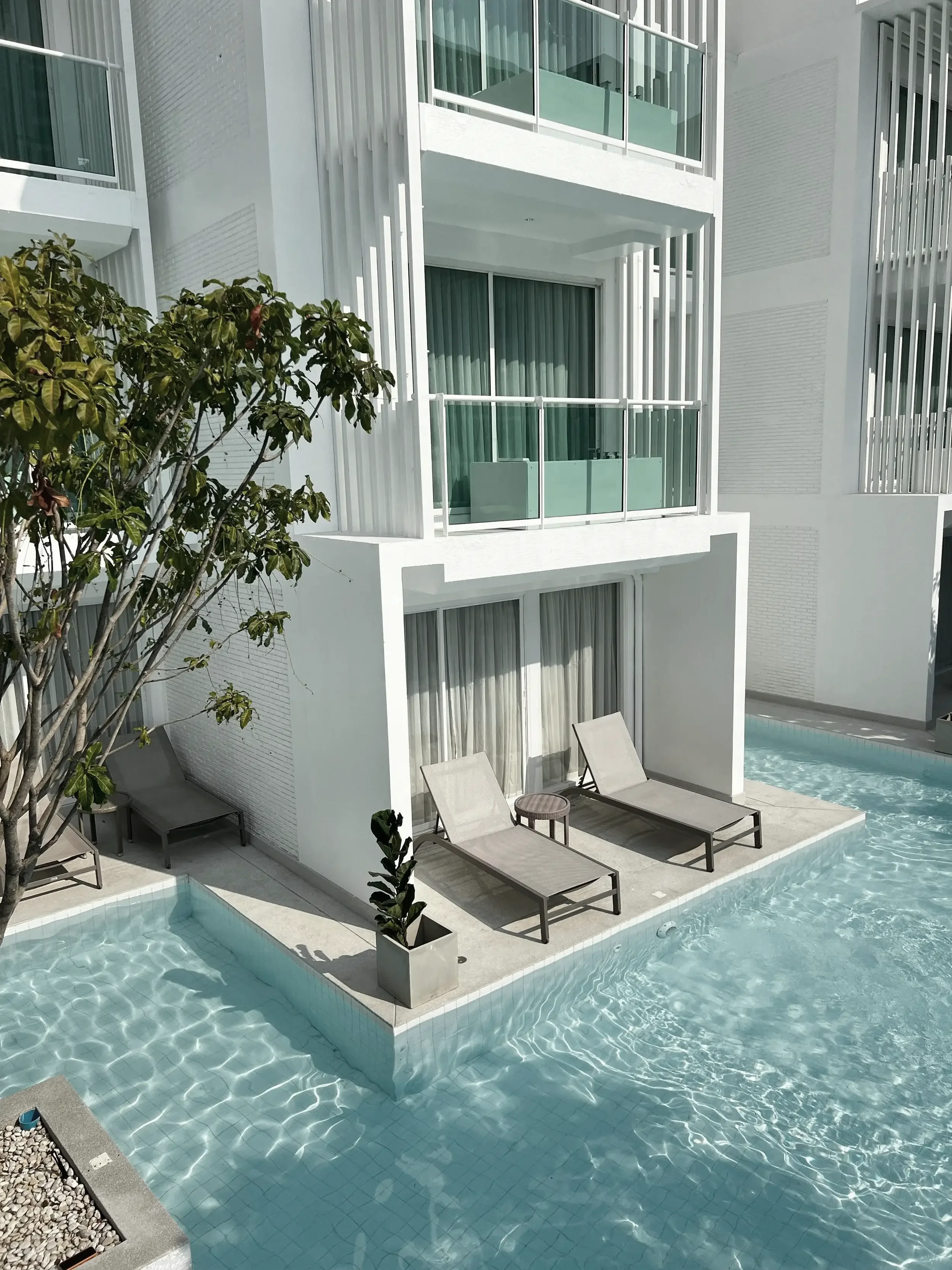 Modern white apartment building with glass balconies, outdoor pool with lounge chairs, and potted plants.