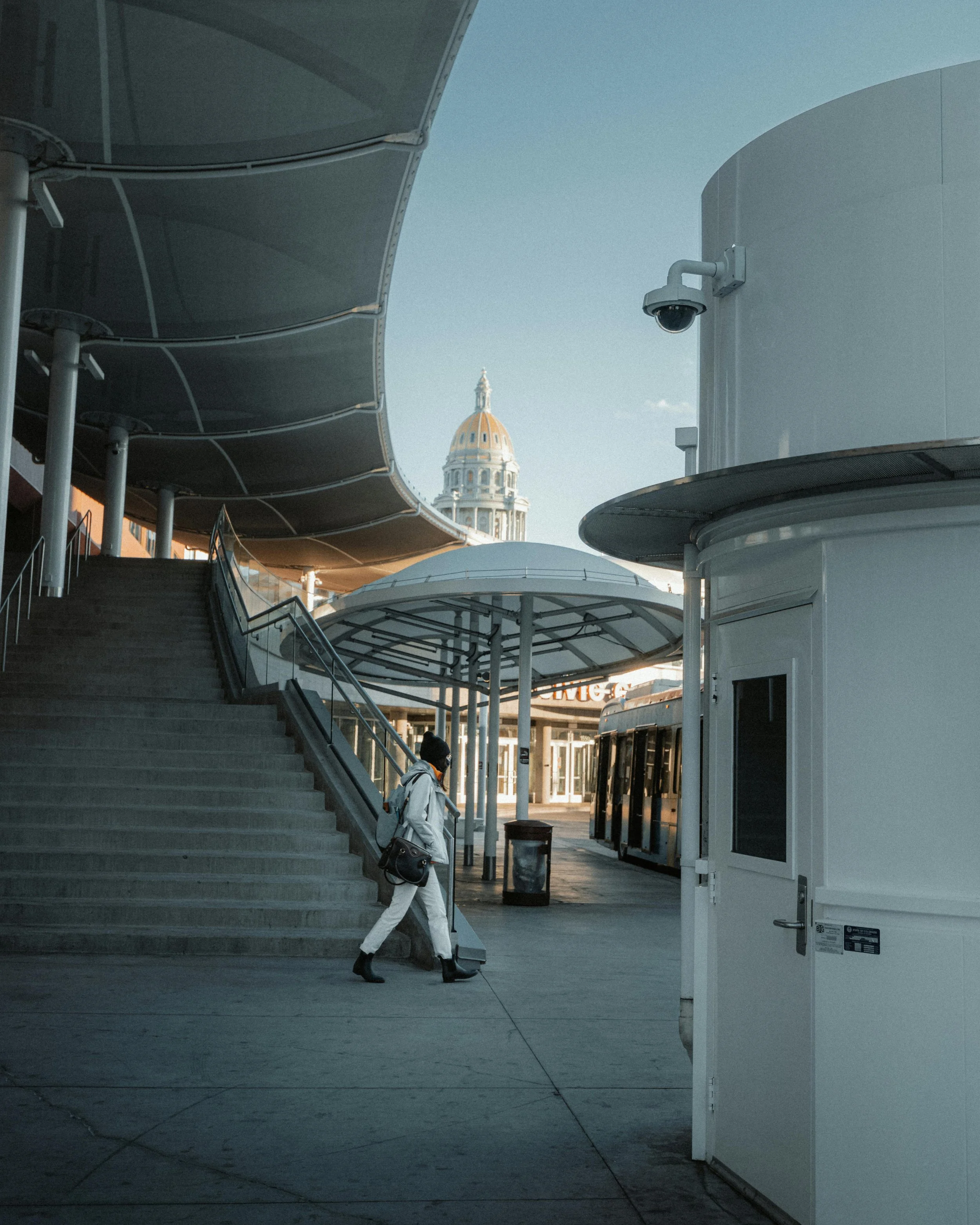 Person walking up stairs outside a modern building with a domed structure and a historic building with a clock tower in the background.