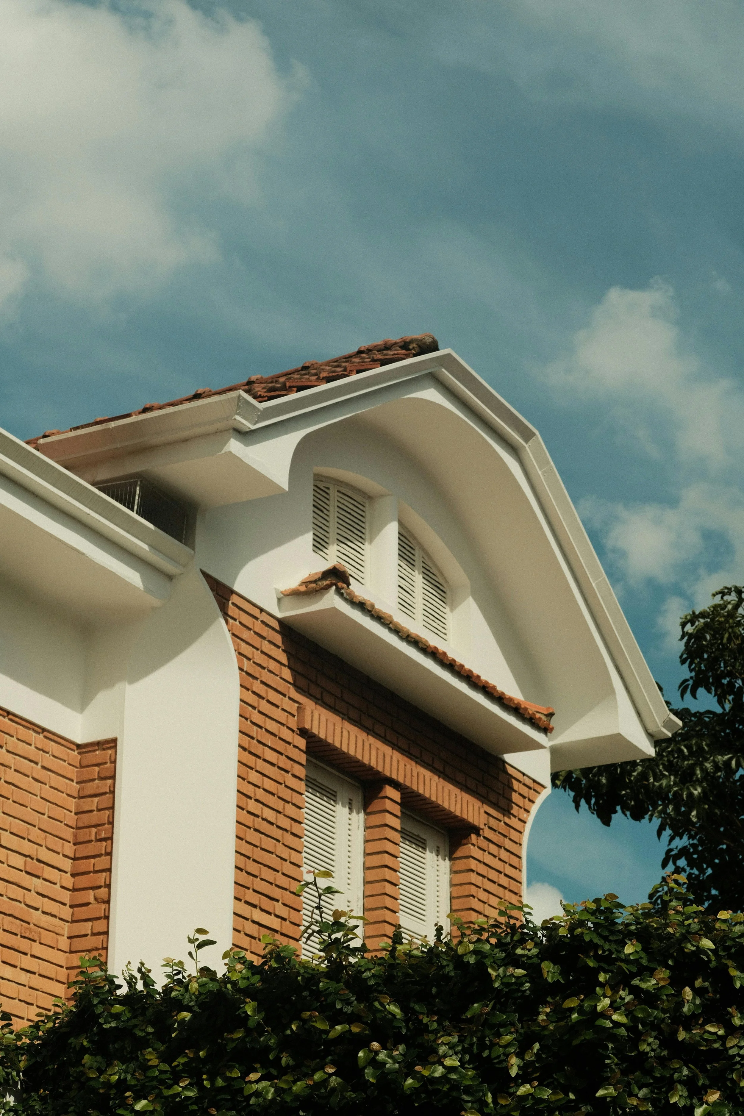 Close-up of a house with red brick and white exterior, showing roof with shingles, arched window, and lower windows with shutters, partly obscured by greenery.
