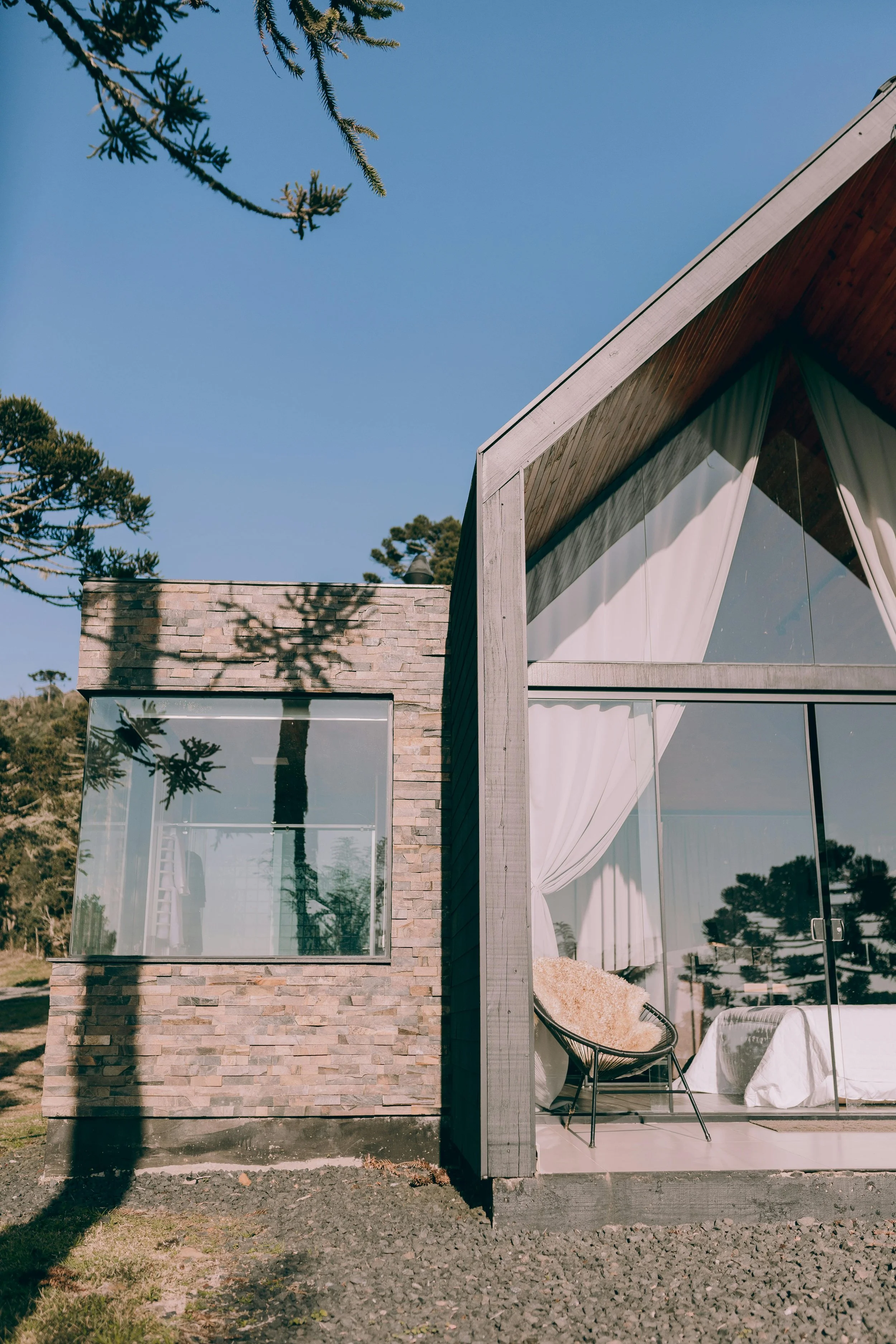 A modern house with large glass windows, wooden exterior, and white curtains, situated outdoors with trees around under a clear blue sky.