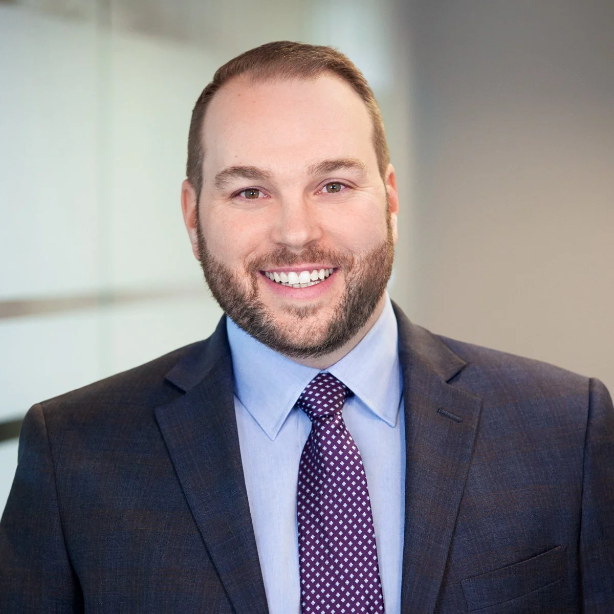 A man with a beard and short brown hair smiling, dressed in a dark suit, light blue shirt, and purple patterned tie, in an office setting.