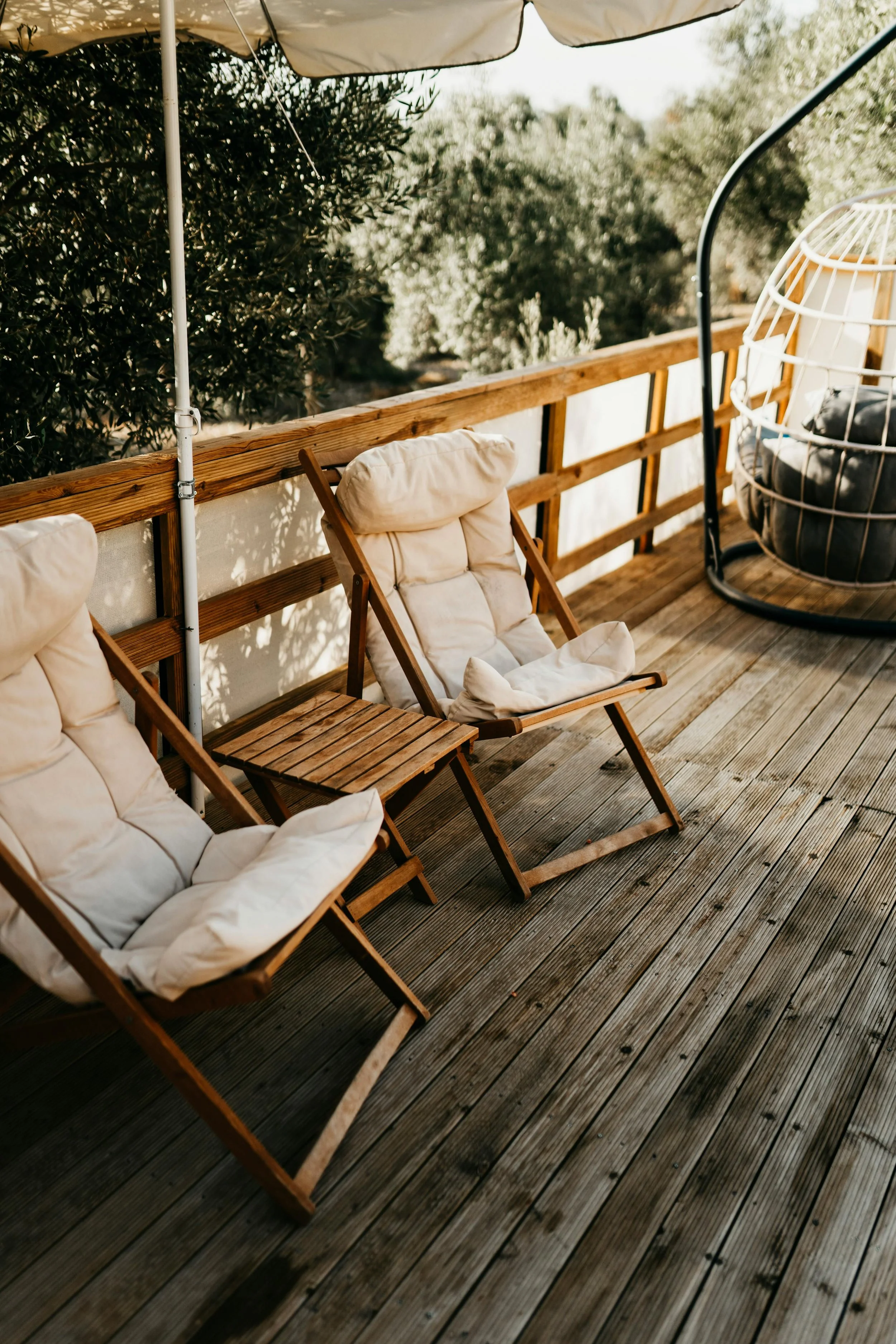Outdoor wooden deck with cushioned wooden chairs, a small wooden side table, a hanging egg chair with cushion, and surrounding greenery.