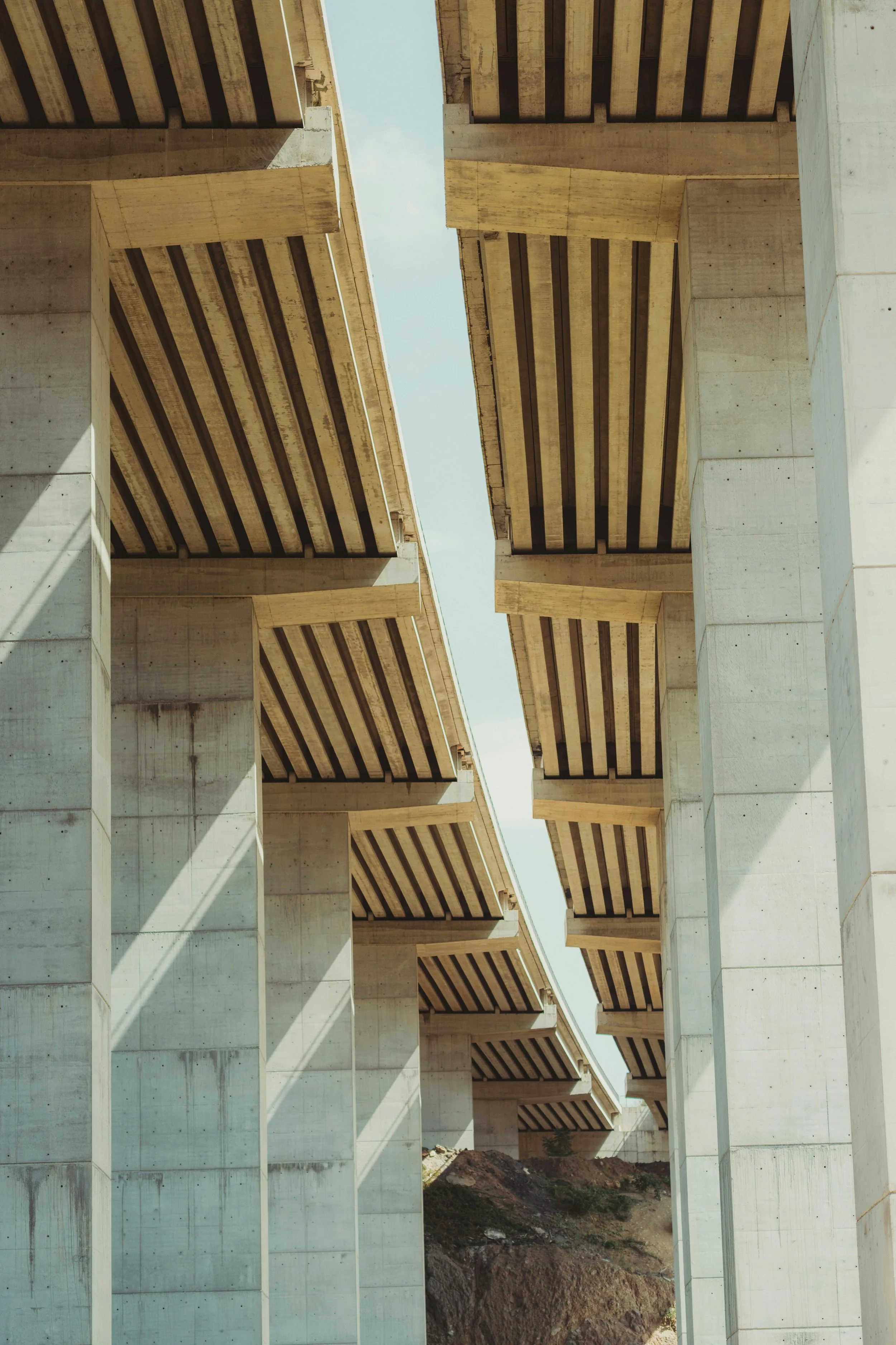 Underneath view of a large concrete bridge with multiple supports and wooden underside beams, with shadows cast on the supports and a partly cloudy sky in the background.