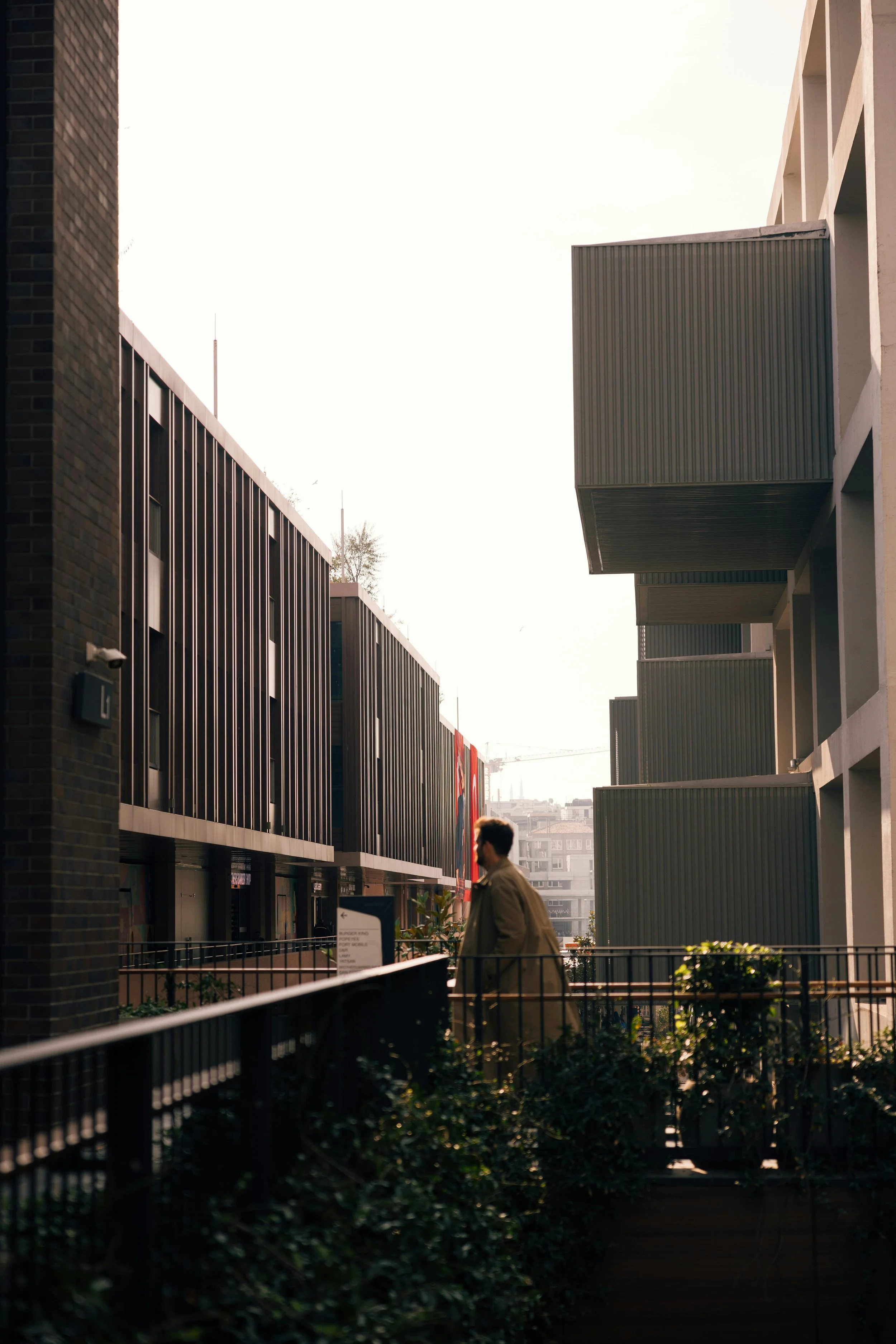 A man walking along a modern urban street with contemporary buildings, greenery, and a railing, during daylight hours.