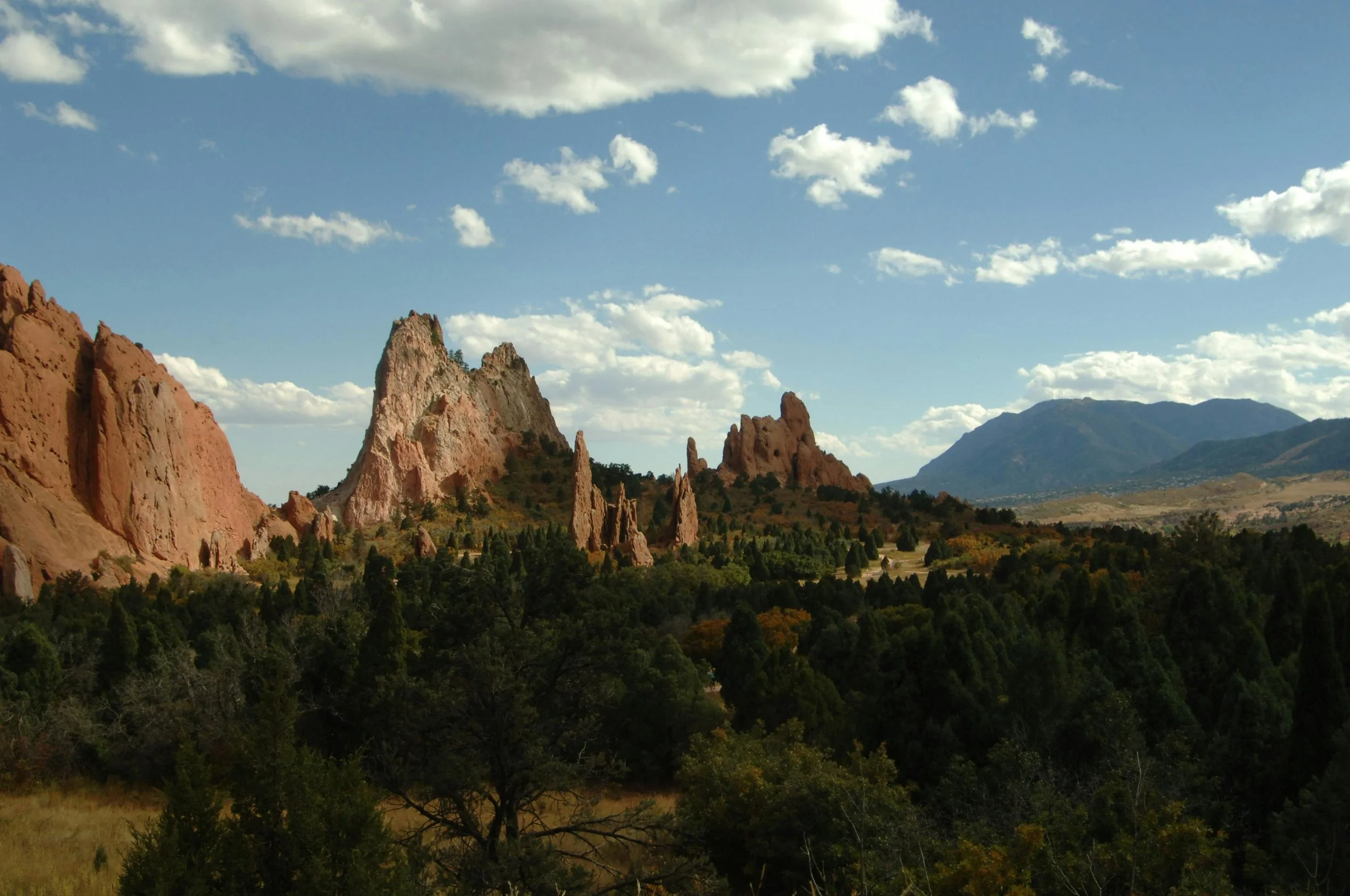 Scenic view of rocky mountains with red and white formations, green trees in the foreground, and a blue sky with scattered clouds.
