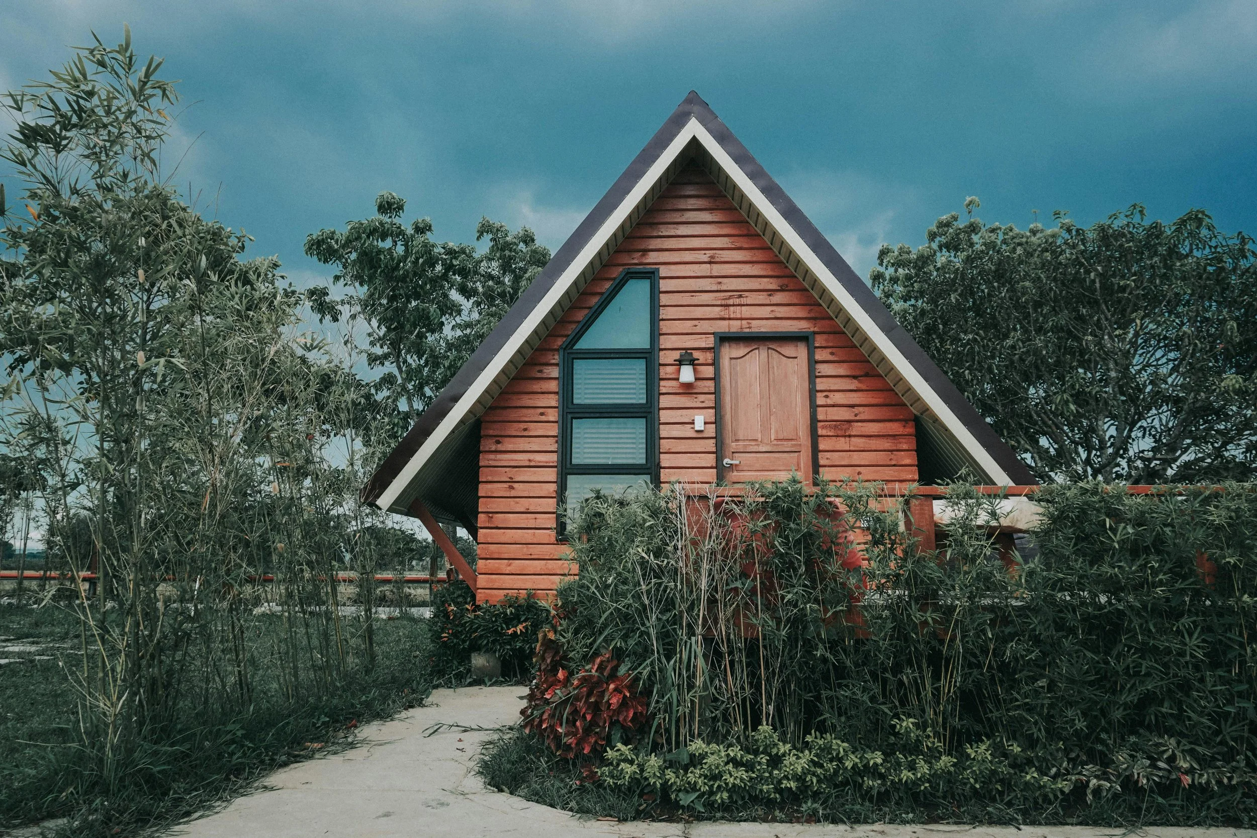 A small A-frame wooden house with a triangular roof surrounded by greenery and trees, under a cloudy sky.