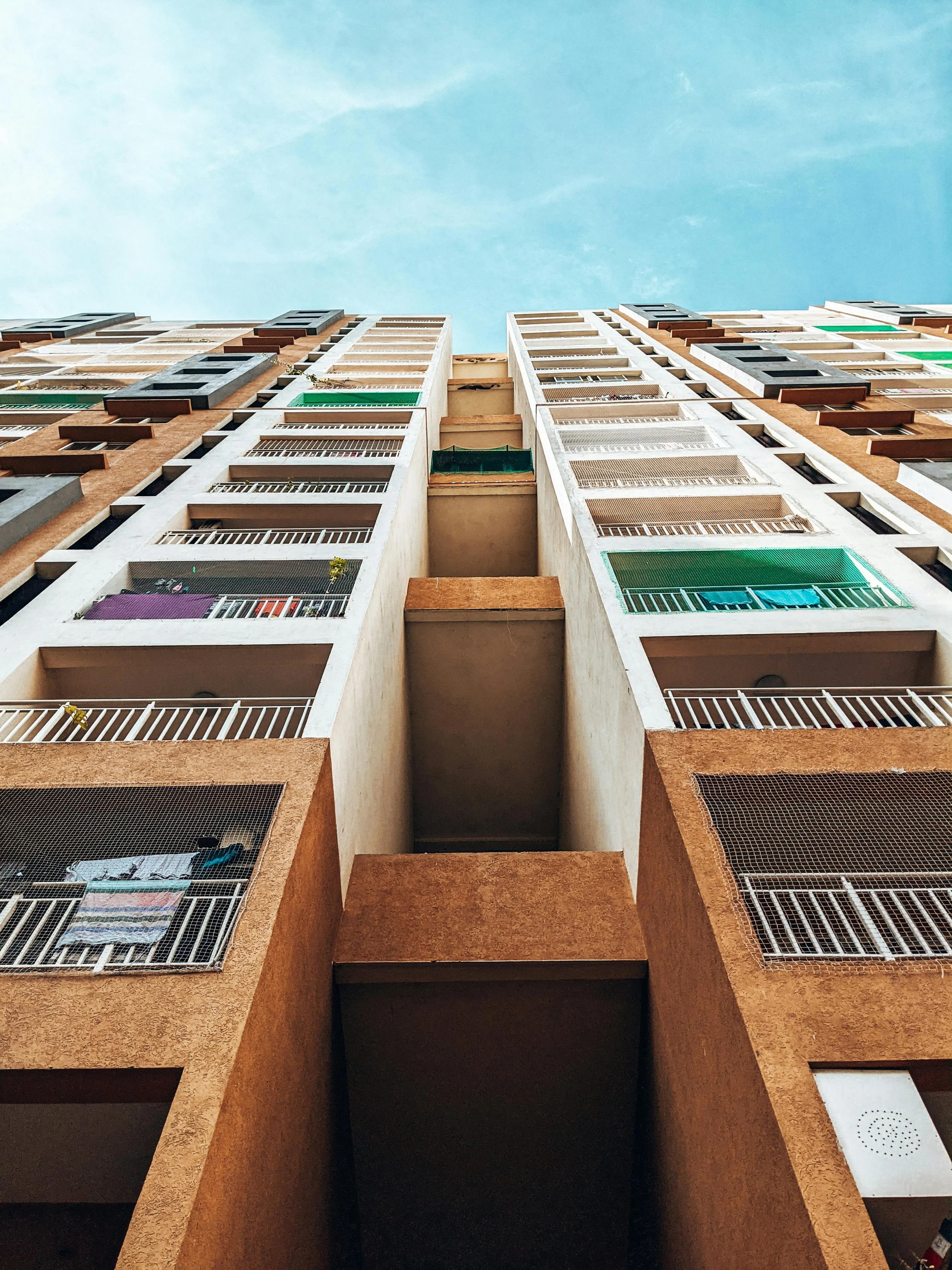Looking up at a tall apartment building with multiple balconies and a clear blue sky.