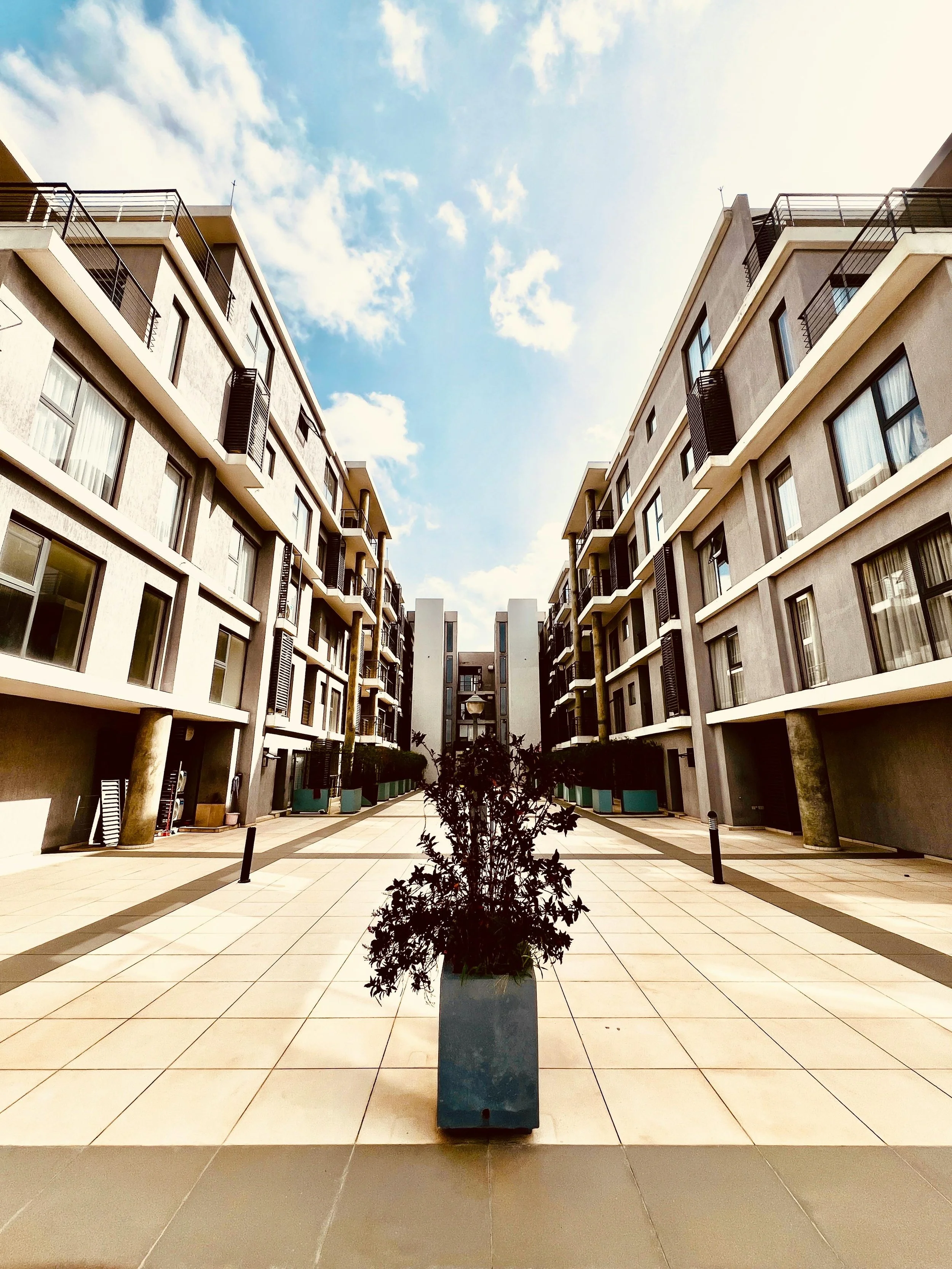 Modern apartment complex with a courtyard, featuring a potted plant in the center and blue sky with clouds overhead.