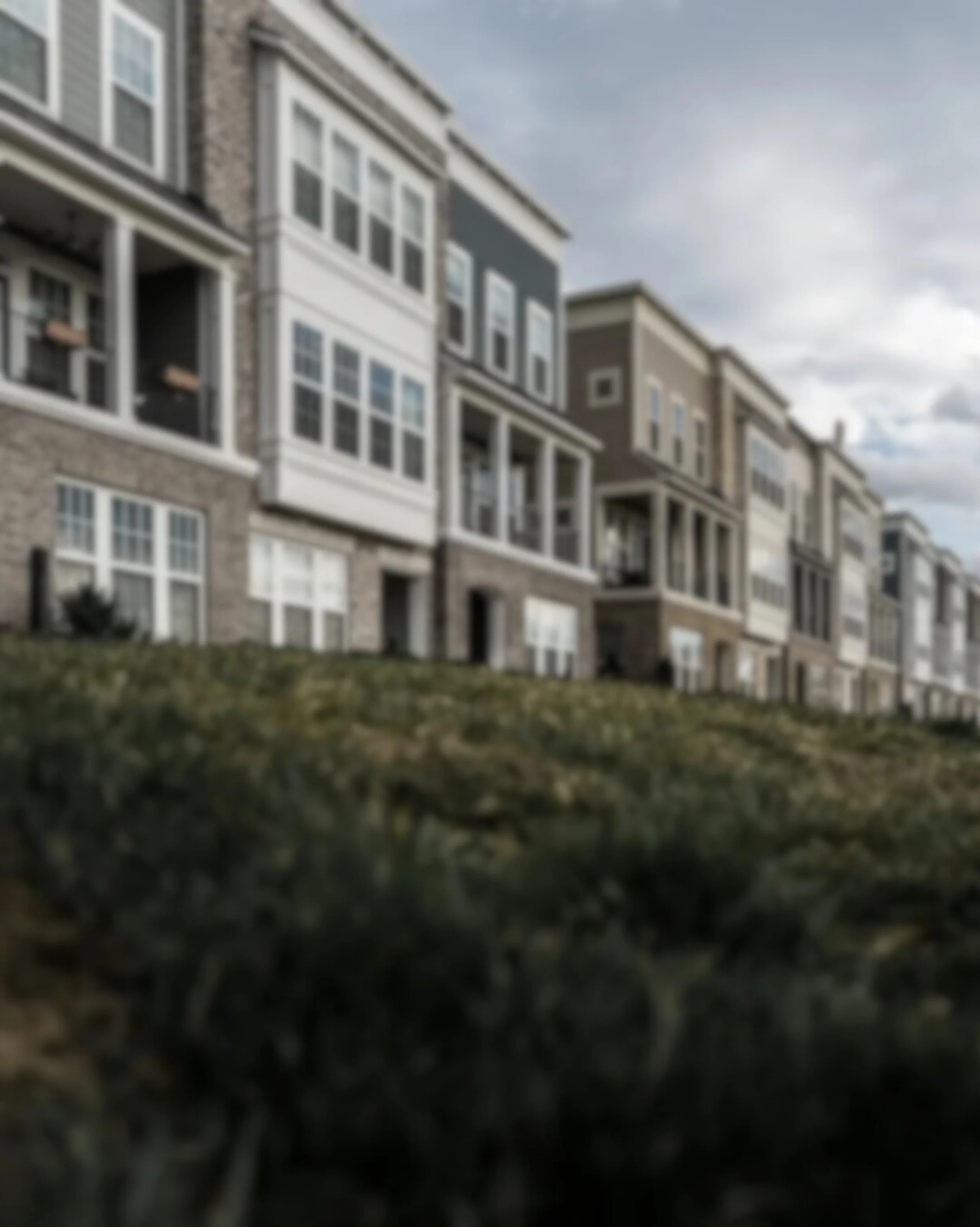 Row of modern multi-story apartment buildings with large windows and balconies, seen from a grassy area in the foreground under a cloudy sky.
