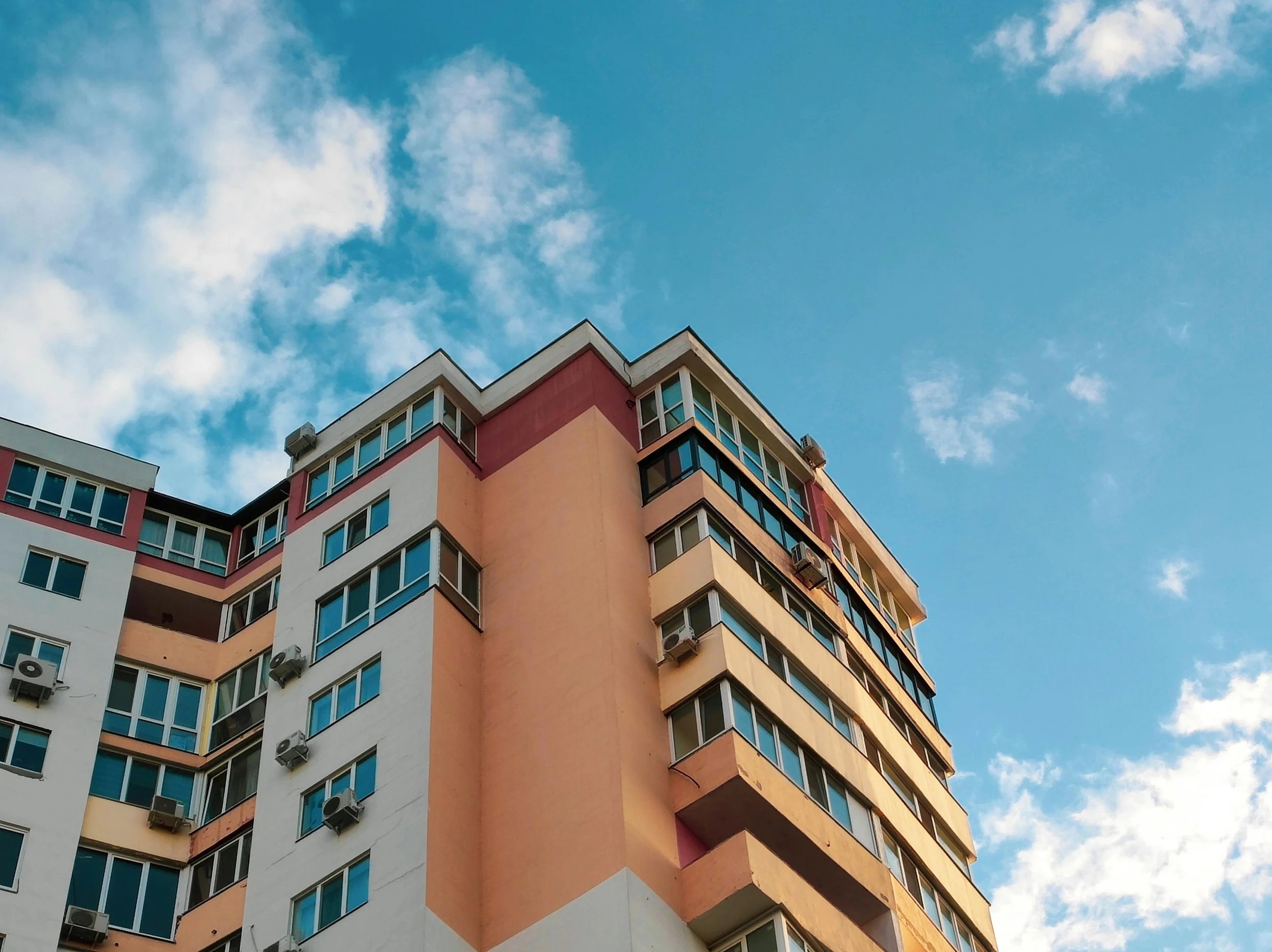 Close-up of a multi-story apartment building with orange, white, and pink exterior walls, multiple windows, and air conditioning units, against a background of a partly cloudy blue sky.