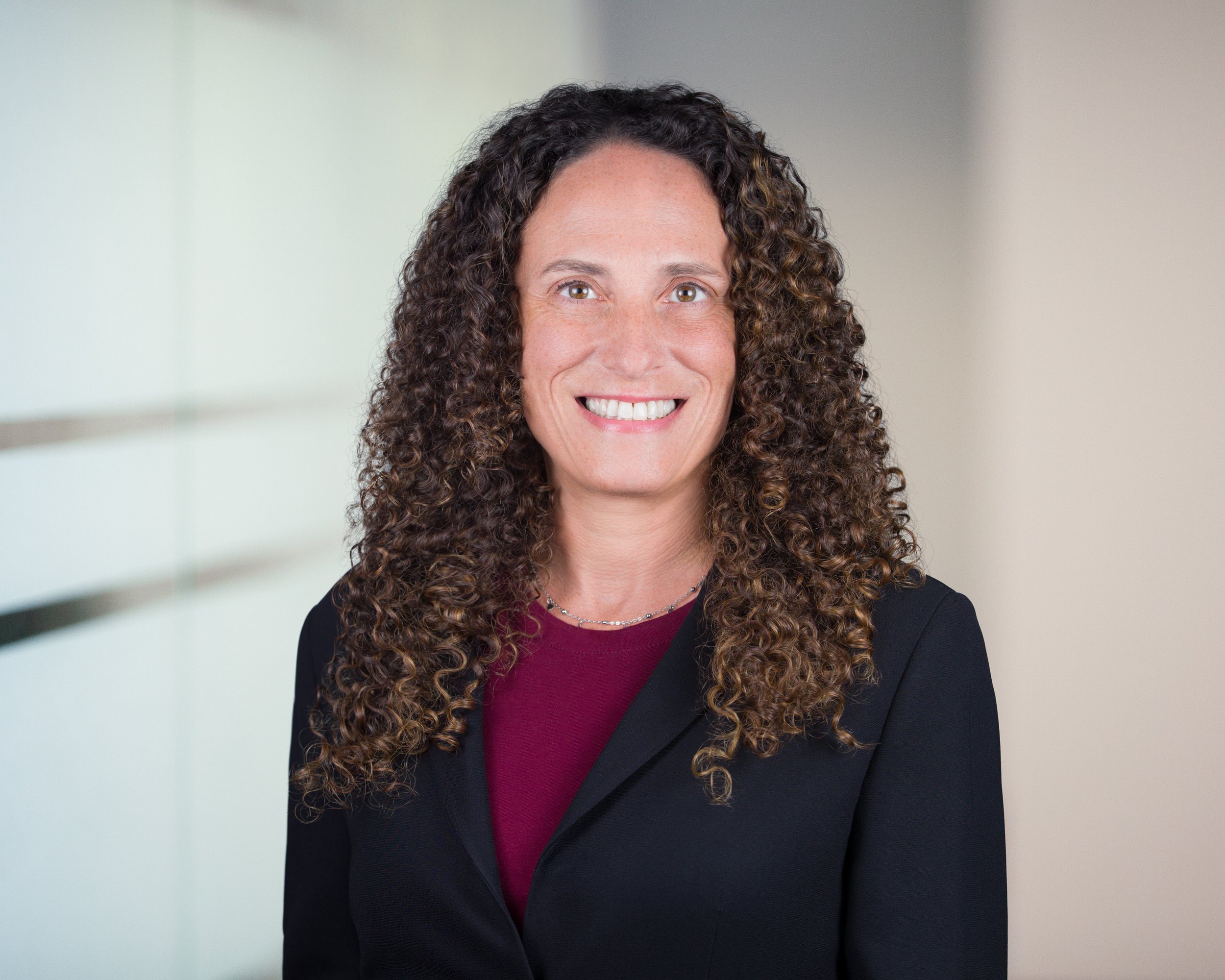 A woman with long curly brown hair, wearing a black blazer and a maroon top, smiling in a professional setting.