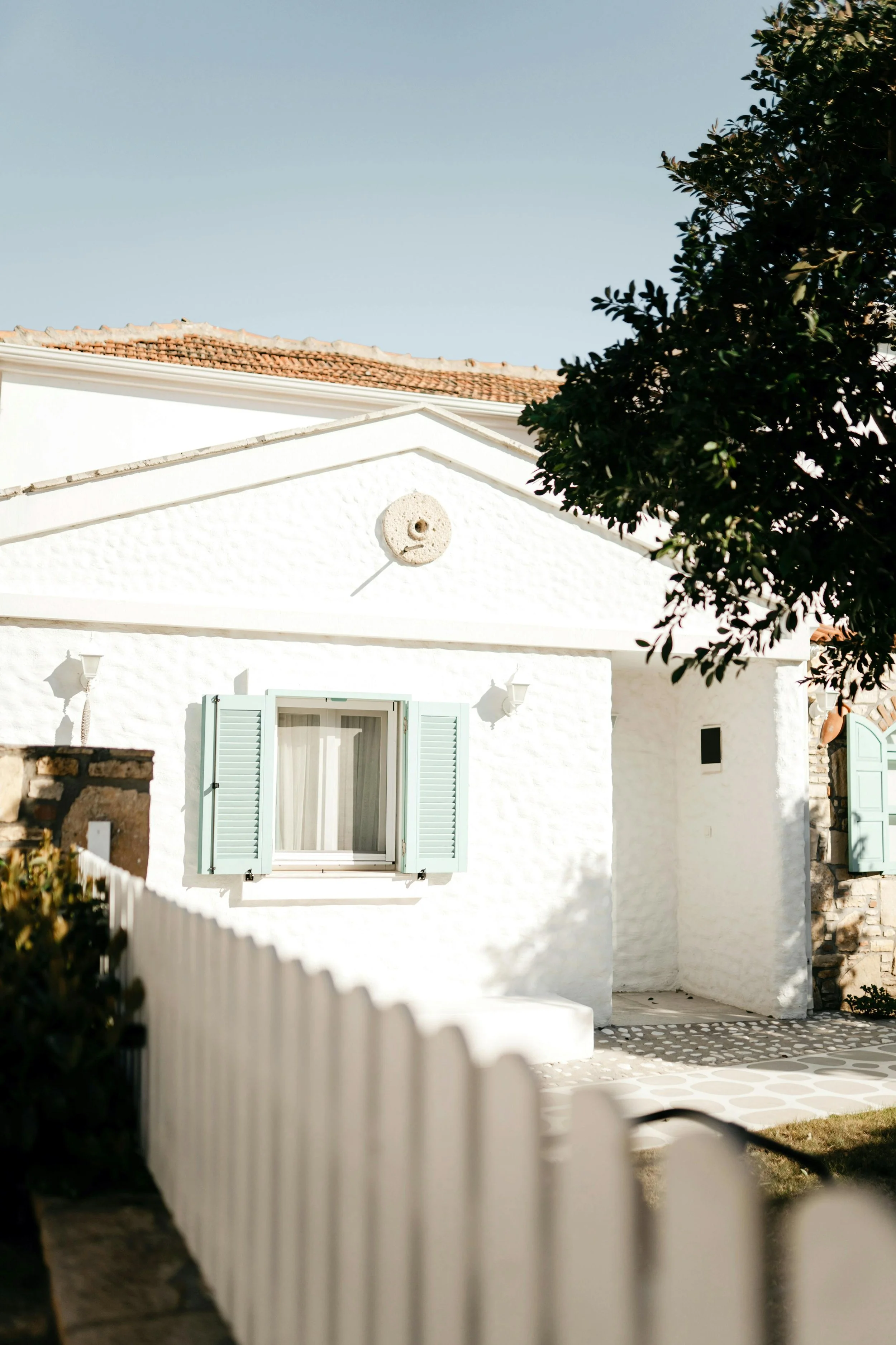 A white house with a tiled roof, blue shutters, and a small front yard enclosed by a white picket fence, under a clear blue sky.
