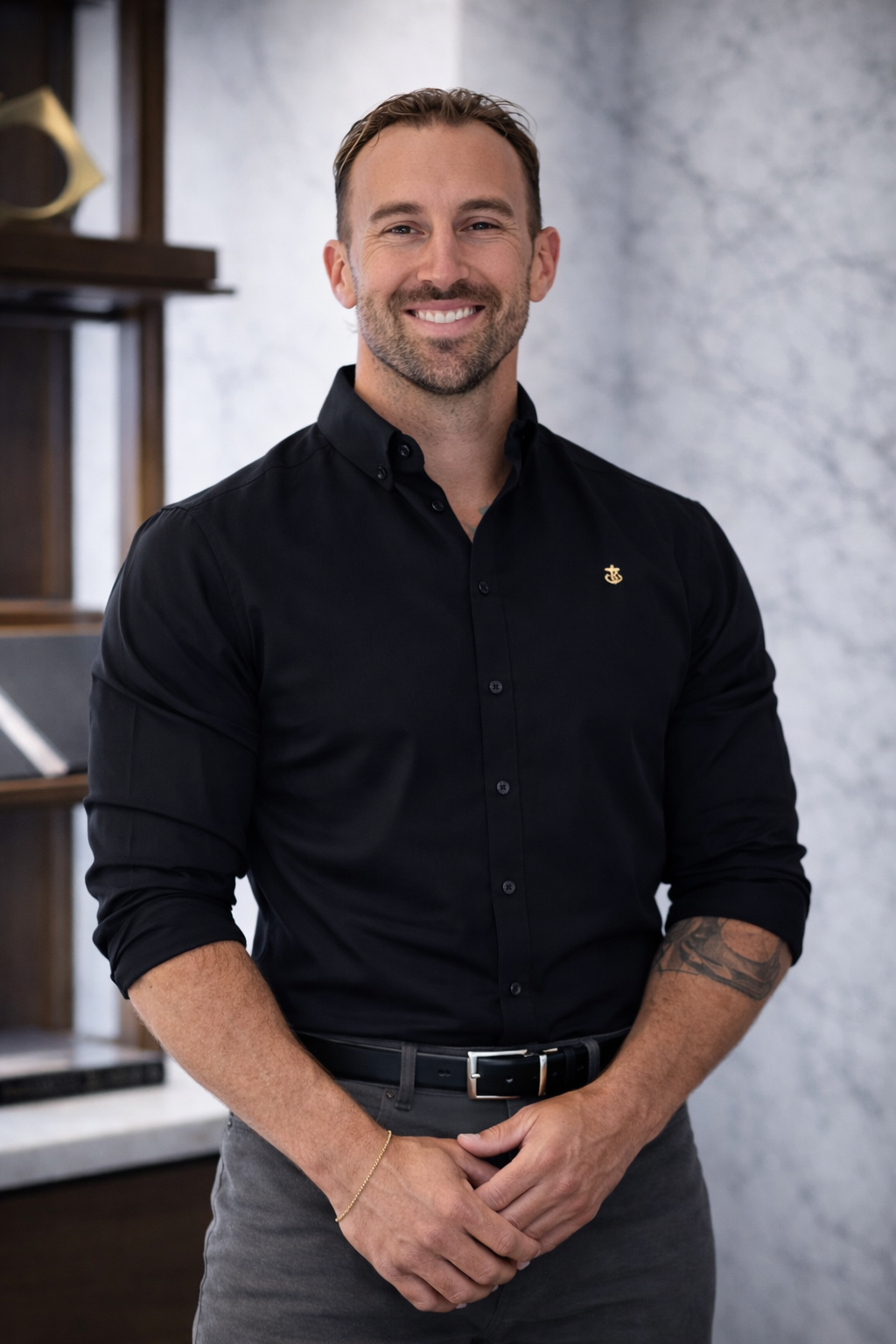 A smiling man with short brown hair and a beard, dressed in a black button-up shirt with rolled-up sleeves, standing indoors against a background of a gray wall and wooden shelves.
