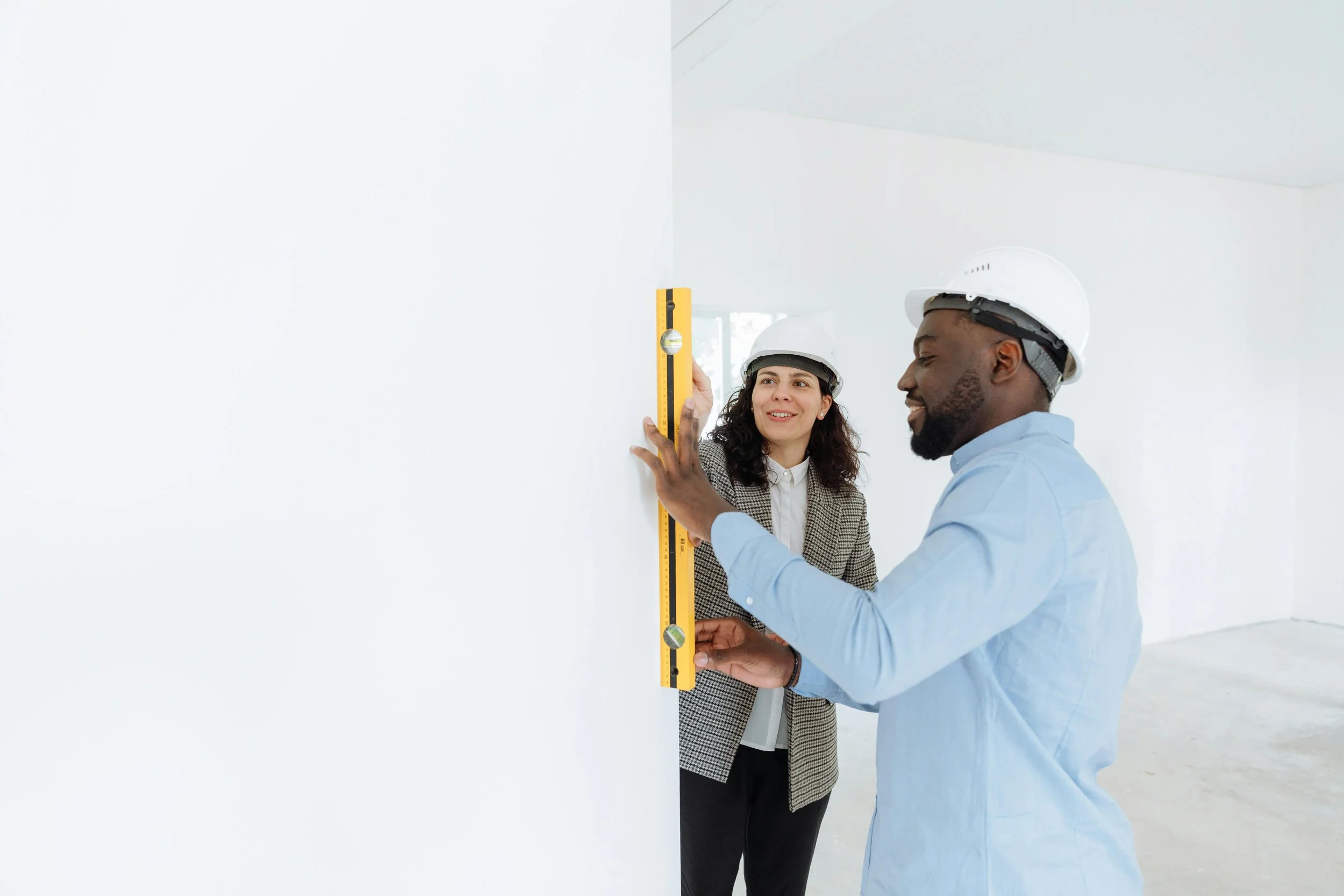 Two construction workers, a woman and a man, wearing white safety helmets, are measuring a wall with a yellow spirit level in a bright, white-room environment.